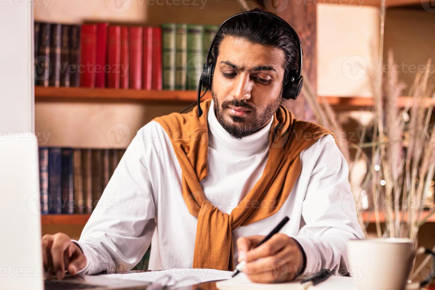Online Teaching. Arab Male Teacher Sitting At Laptop Wearing Headset And Taking Notes Having Remote Distant Class At Home. Distance Study, Webinar And E-Teaching Concept. photo