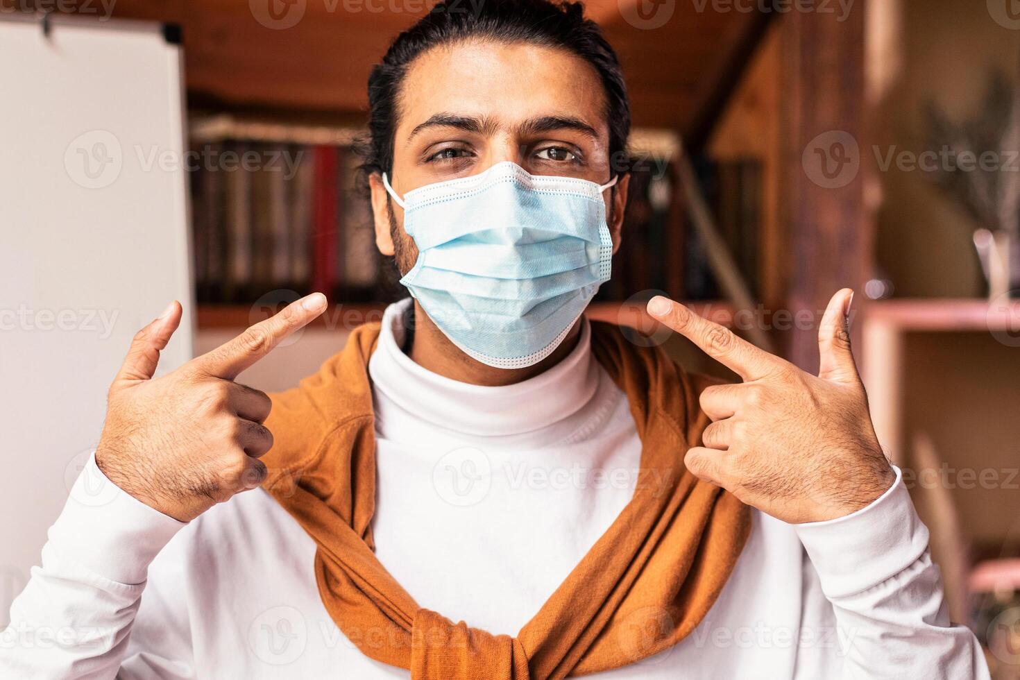 Covid quarantine at school. Indian teacher pointing at face mask looking at camera standing near blackboard indoors, announcing remote learning online due to coronavirus pandemic outbreak photo
