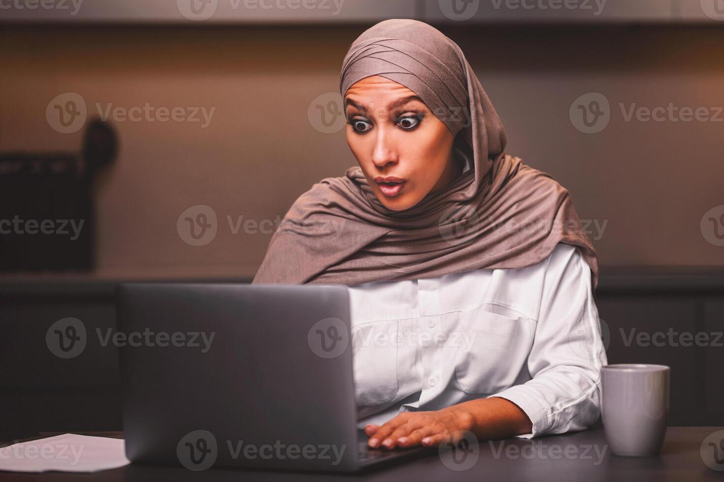 Shocked Muslim Woman Looking At Laptop Computer Browsing Internet And Reading Shocking News Online Sitting In Modern Kitchen At Home, Wearing Hijab. Omg Concept photo