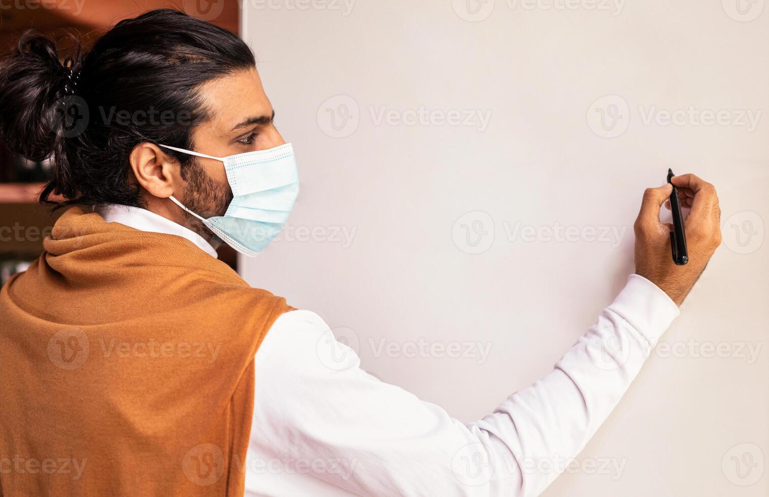 Teacher Man In Face Mask Teaching Writing On Blank White Blackboard Having Class Standing Indoors. Safe School And University Education, Tutoring, Distant And Online E-Teaching Concept. Mockup photo
