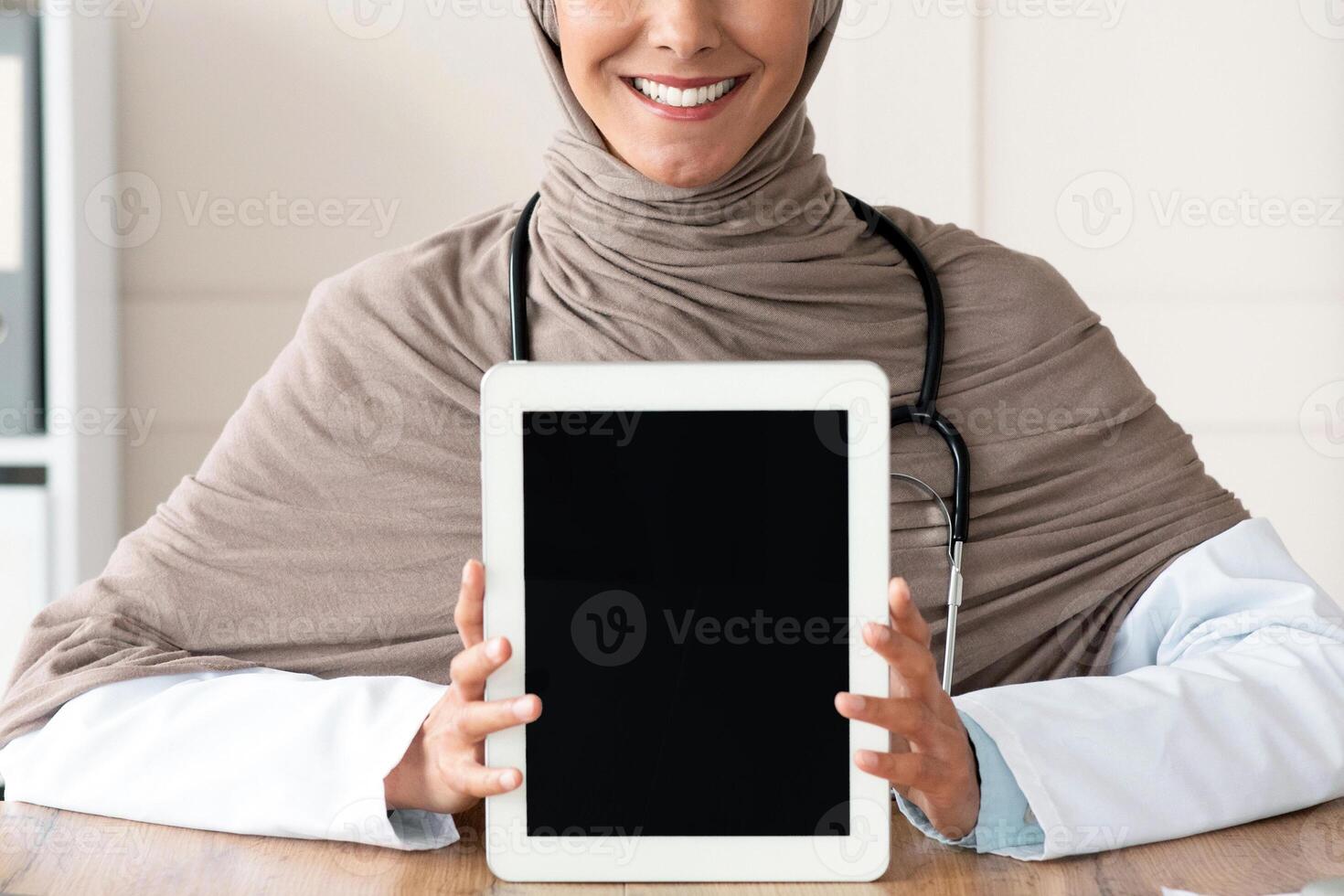 Cheerful female doctor muslim woman in hijab showing digital tablet with blank screen, mockup, copy space. Smiling doctor using modern technologies in her job, working and training online photo