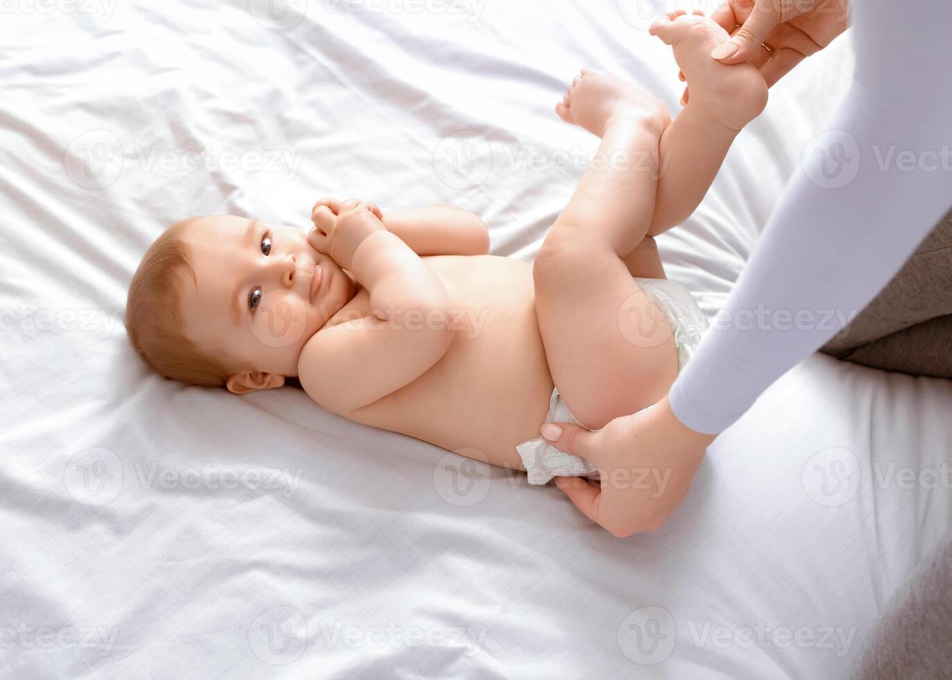 Unrecognizable mother changing her smiling little baby's nappy on white, top view. Cute toddler blonde boy having fun while his mom dressing him up, playing with his feet, closeup, empty space. photo