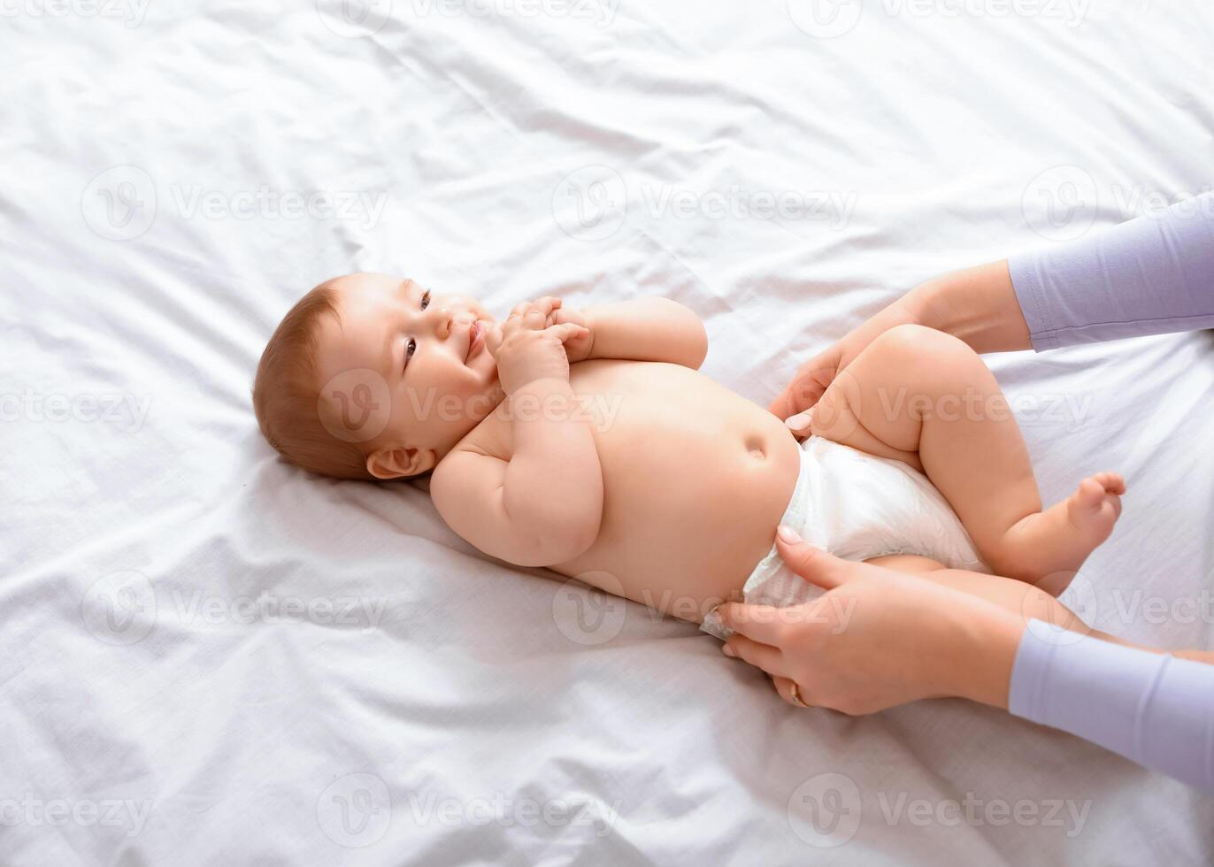 Unrecognizable mother changing her smiling little baby's diaper on white, top view. Cute toddler blonde boy having fun while his mom dressing him up, playing with his fingers, closeup, empty space. photo