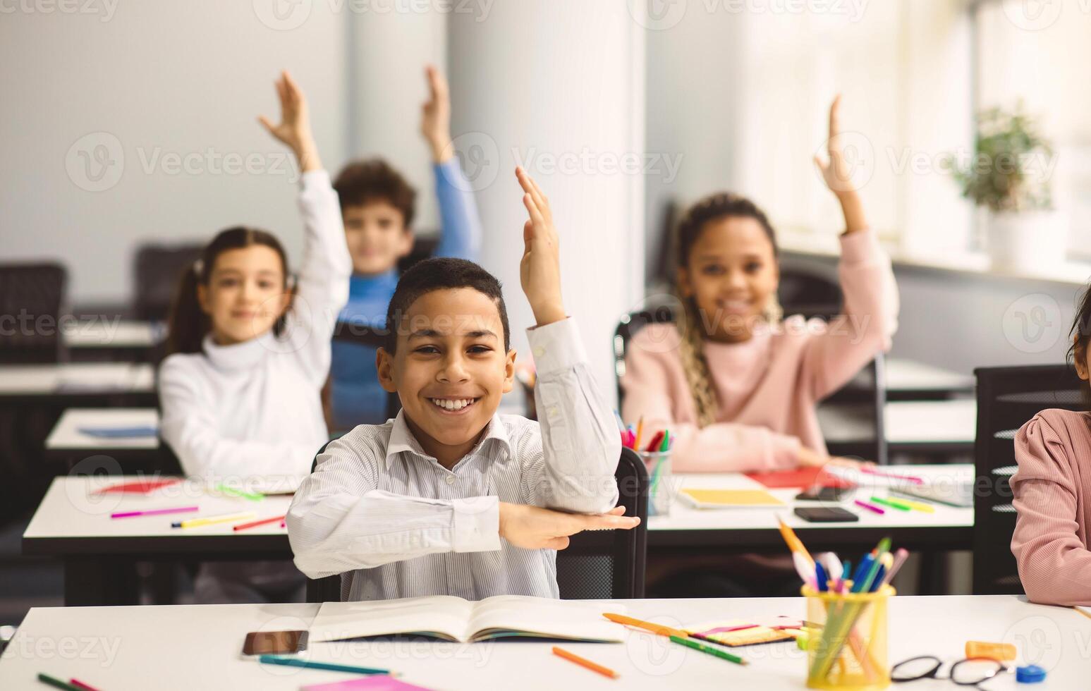 School Reopening, Back To Normal Lifestyle Concept. Portrait of excited little schoolboy raising hand up in the air to answer a question with all his classmates in blurred background, selective focus photo