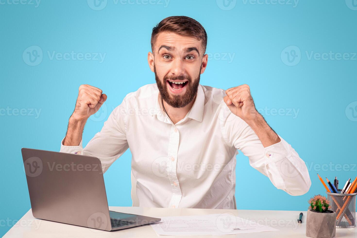 Positive young businessman making YES gesture while working on laptop at desk over blue studio background. Professional office manager celebrating achievement or success at workplace photo