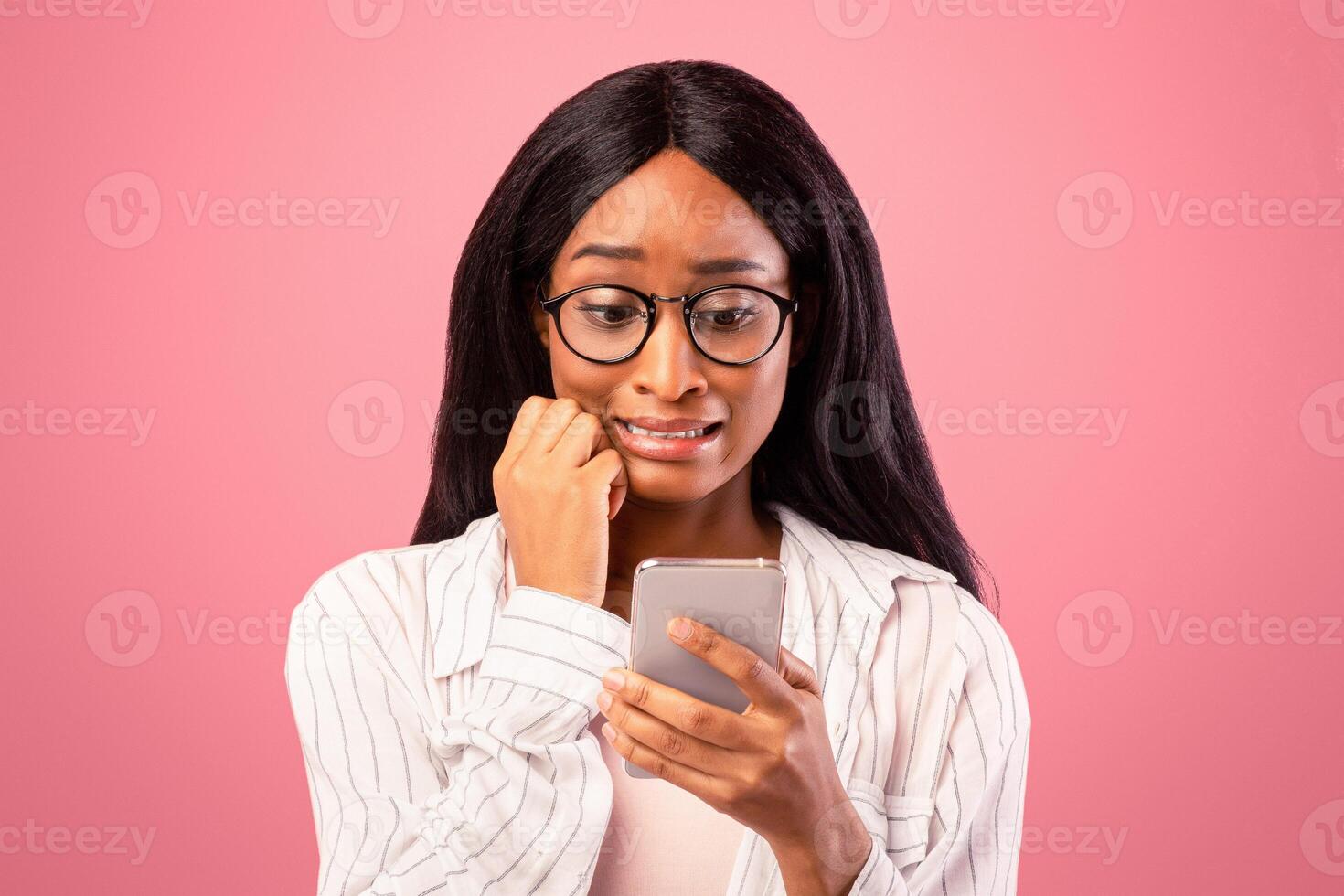 Oops. Embarassed black woman looking at mobile phone screen in confusion on pink studio background. Emotional African American lady making mistake while working in web photo