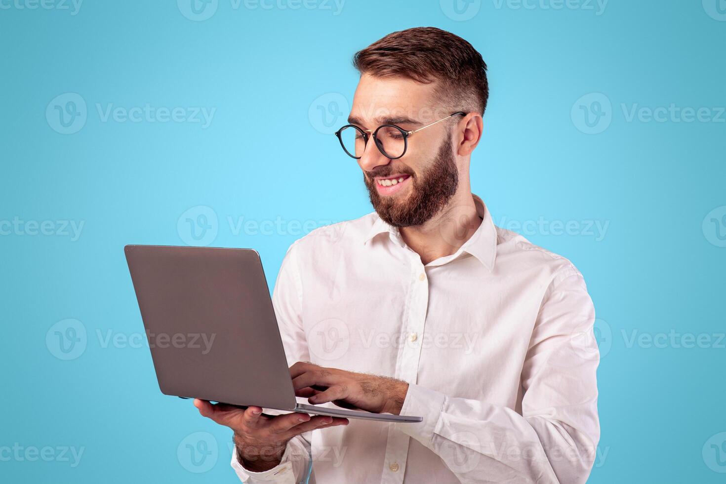 Successful young guy in formal wear using laptop computer over blue studio background. Cheerful businessman having online job, participating in web conference, communicating via internet photo