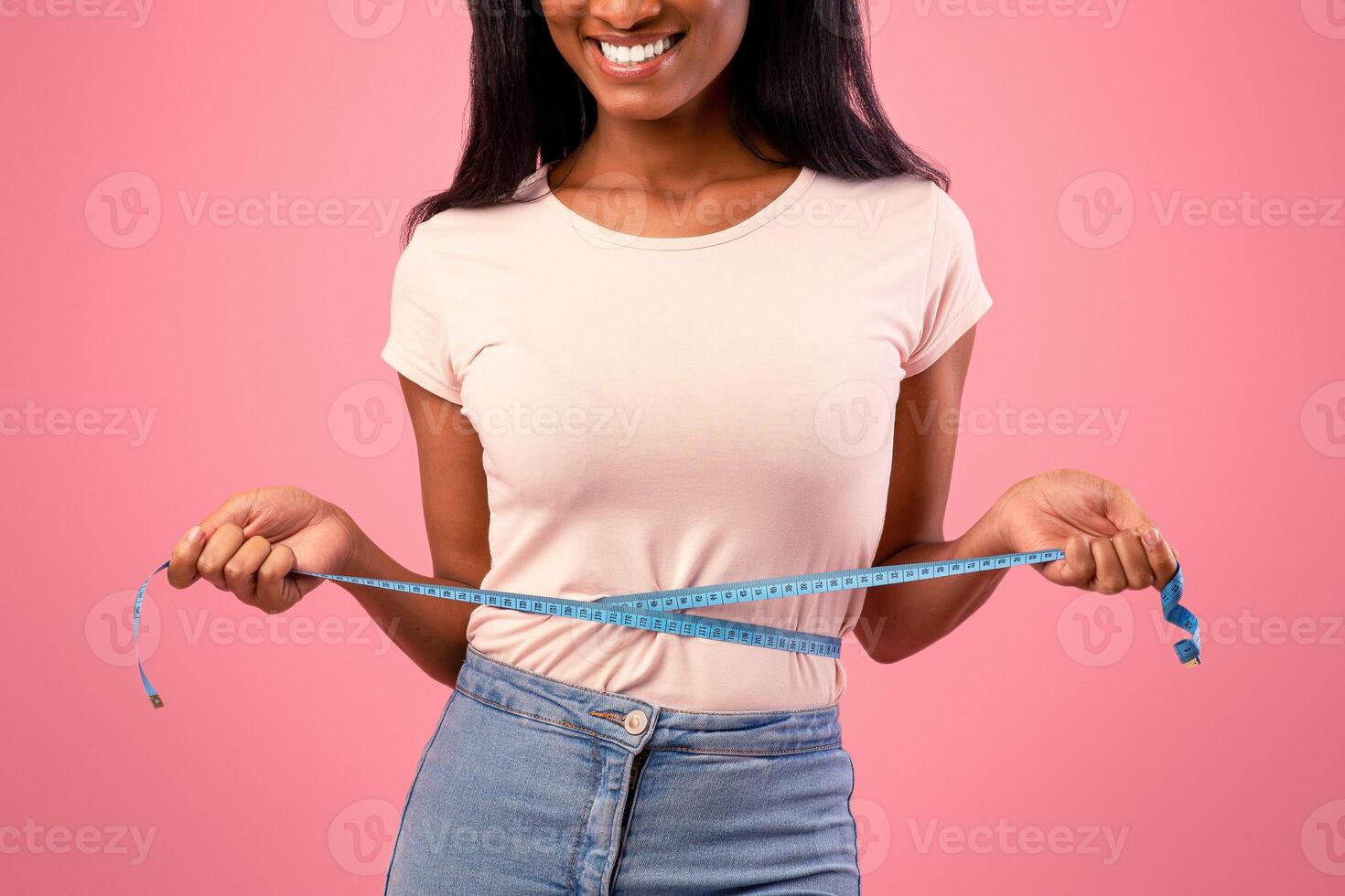 Slimming and dieting concept. Unrecognizable black woman measuring her waist during weight loss over pink studio background. Cropped view of young lady checking her body parameters photo