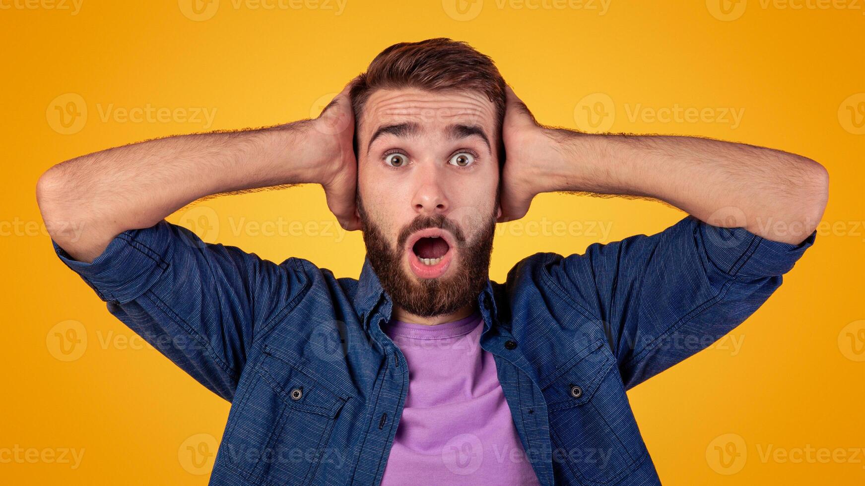 Surprised Caucasian man opening mouth in amazement, looking shocked and stunned on orange studio background. Millennial young guy holding his head in amusement, shouting OMG photo