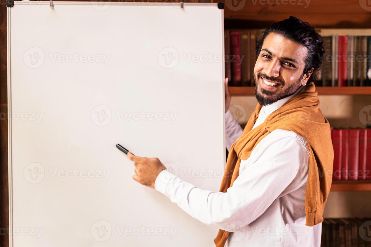 Remote modern education. Indian teacher man teaching online pointing to blackboard explaining English grammar rules standing indoors, smiling to camera. Guy teaches language distantly photo