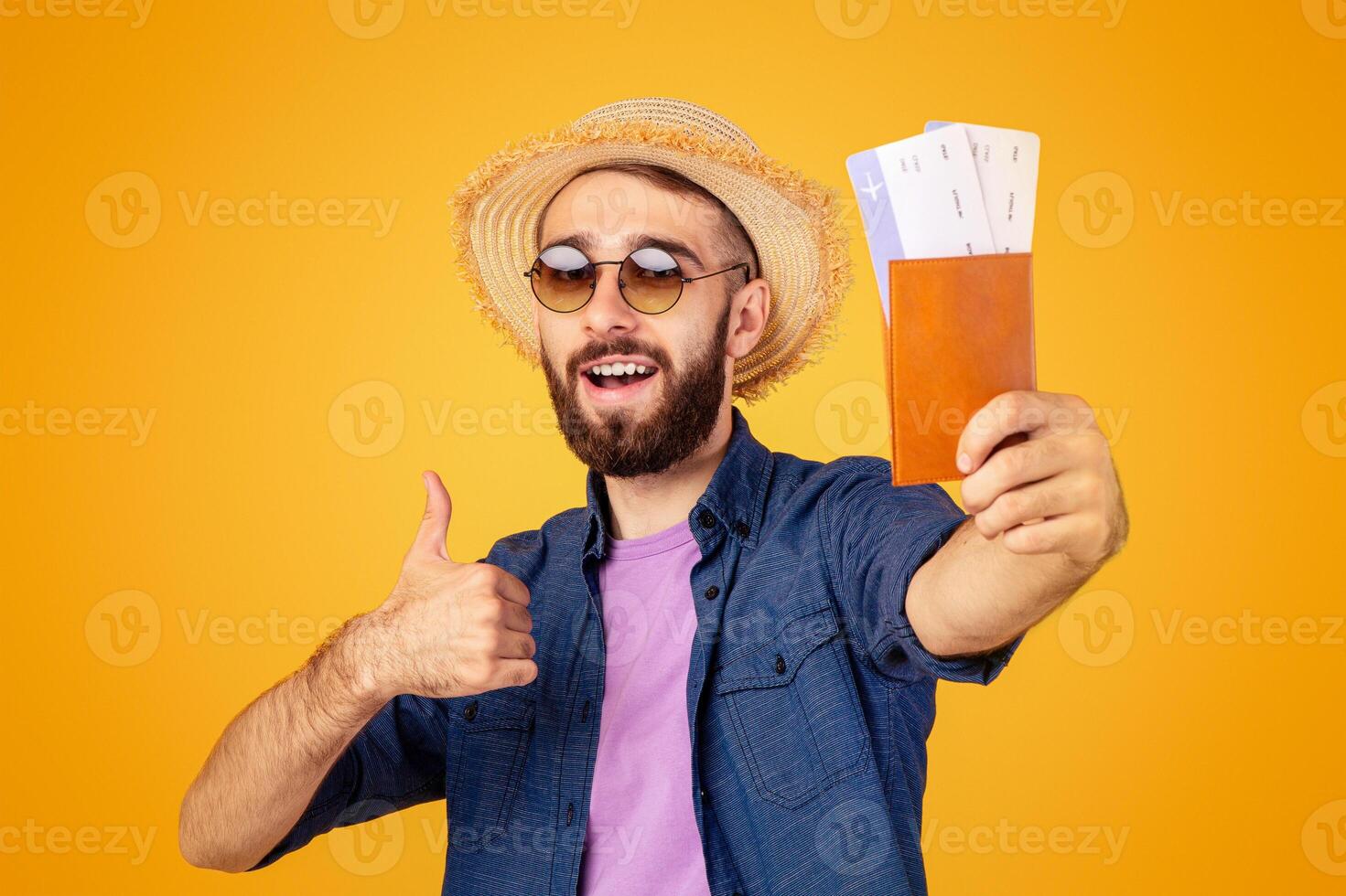Overseas trip. Millennial guy holding international passport with airplane tickets and showing thumb up gesture over orange studio background. Cool tourist with documents ready to travel abroad photo