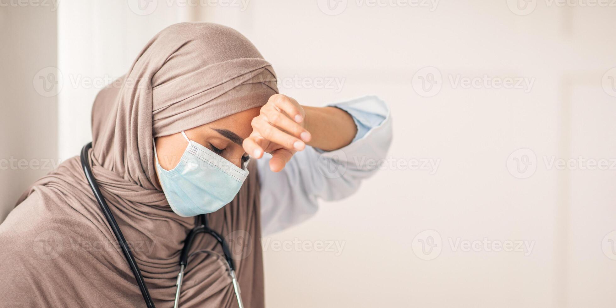 Stressed muslim woman doctor in protective face mask touching her sweating forehead, feeling exhausted after difficult working day, sitting by wall at clinic, copy space. Doctors and COVID-19 pandemic photo