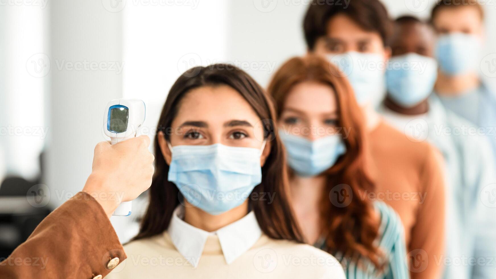 Woman Doing Temperature Screening Measuring Fever For Diverse People Standing In Queue, Using Infrared Contactless Themometer Indoors. Coronavirus, Covid-19 Pandemic Outbreak Concept photo