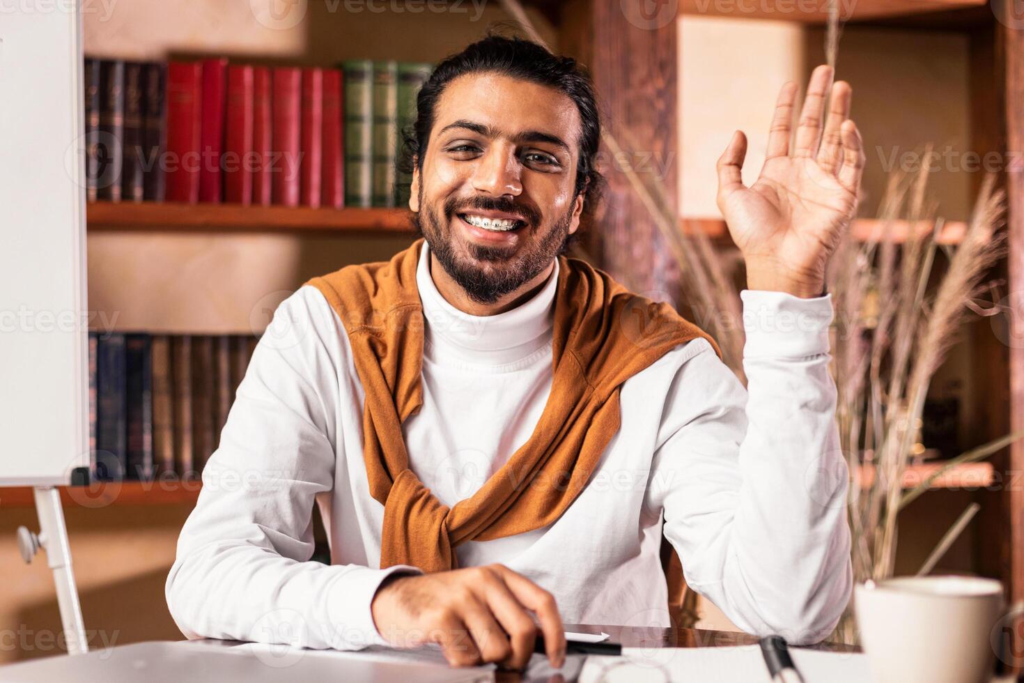 Joyful Indian Teacher Man Waving Hello To Camera Posing Sitting In Classroom With Blackboard Having Online Class Via Call. Distant Learning And Teaching, Virtual Lecture And Webinar Concept photo