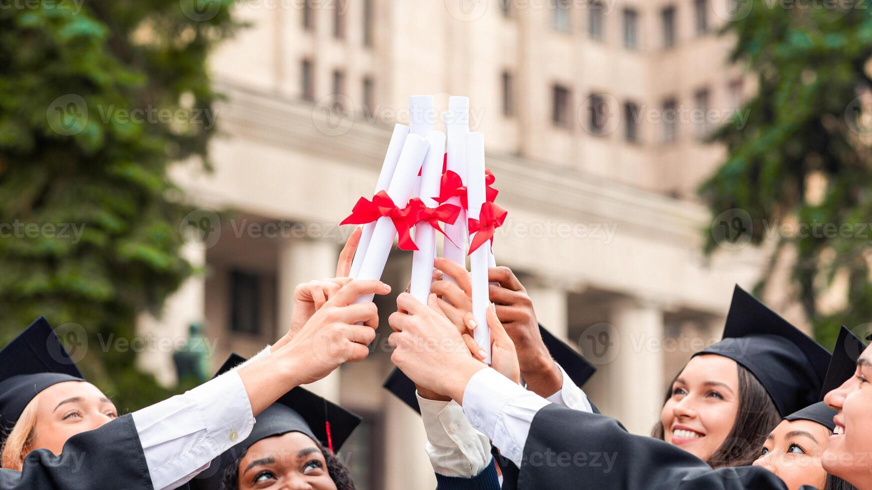 Happy multiracial group of students in graduation caps and robes cheerfully posing at university campus, joining together their diplomas, celebrating graduation, closeup, copy space photo