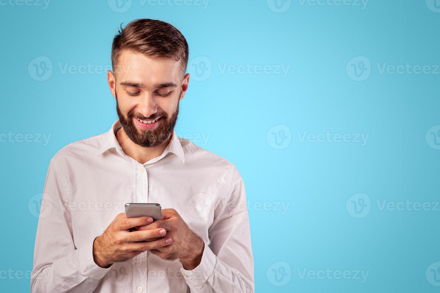 Joyful young man looking at smartphone screen, browsing internet, playing online game, doing shopping on blue studio background. Happy Caucasian guy using modern mobile device photo
