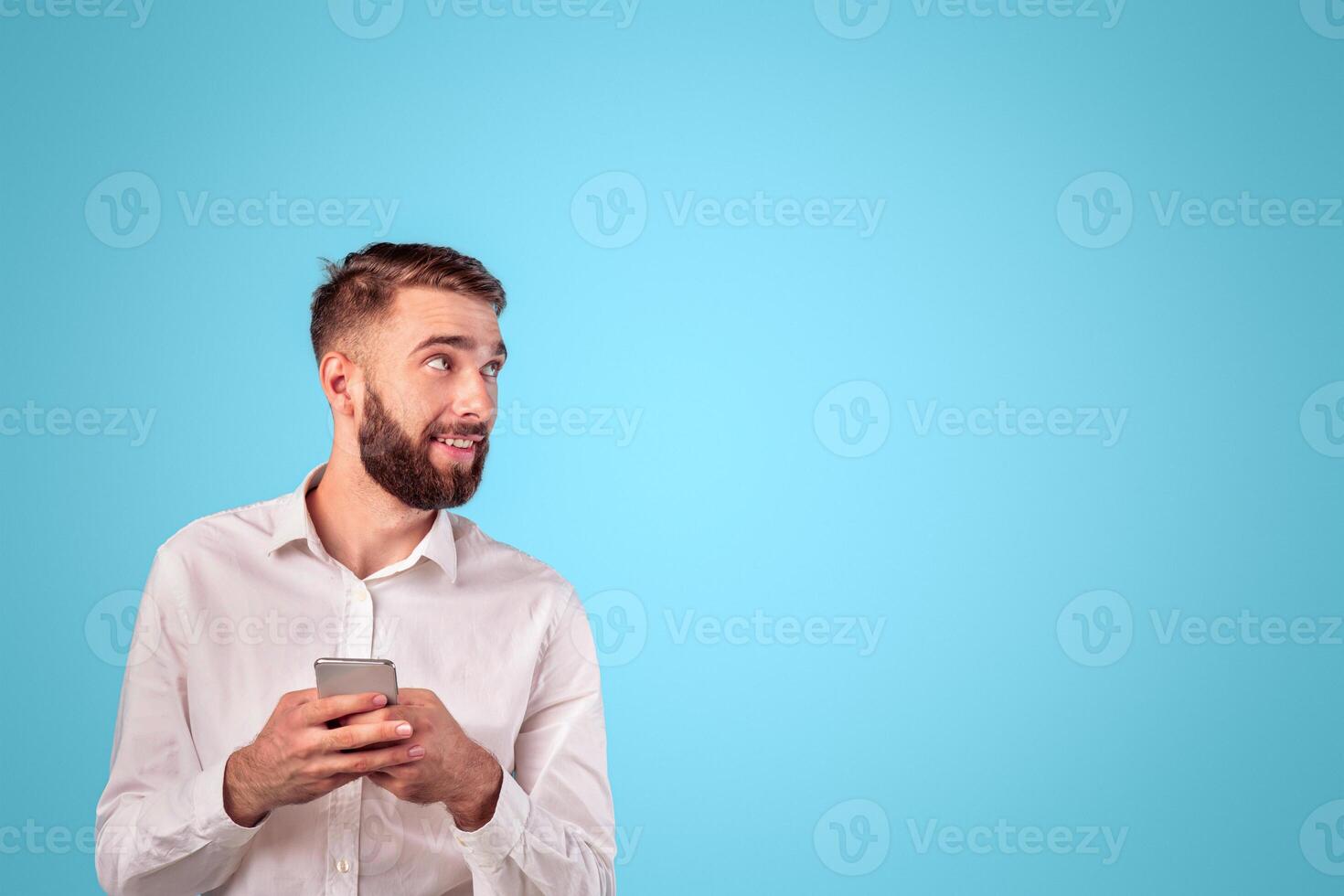 Happy young businessman with mobile phone looking at empty space on blue studio background. Smiling Caucasian guy using modern device for online work or studies, surfing web, checking email photo