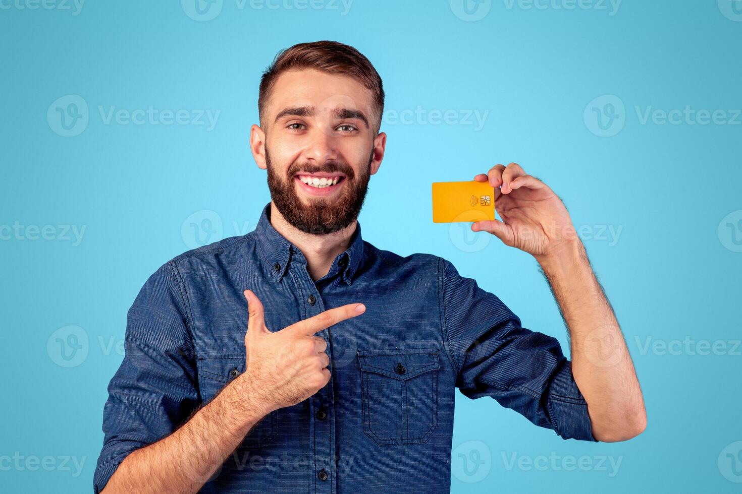 Joyful millennial man pointing at credit card on blue studio background. Happy bank client promoting financial services, using contactless payment. Electronic money concept photo