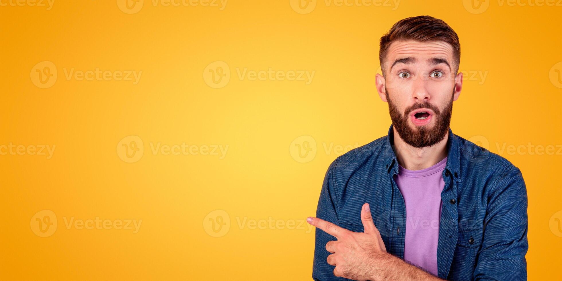 Shocked millennial guy pointing aside at empty space on orange studio background, panorama. Bearded hipster guy advertising your product, offering huge sale or promo, banner design photo