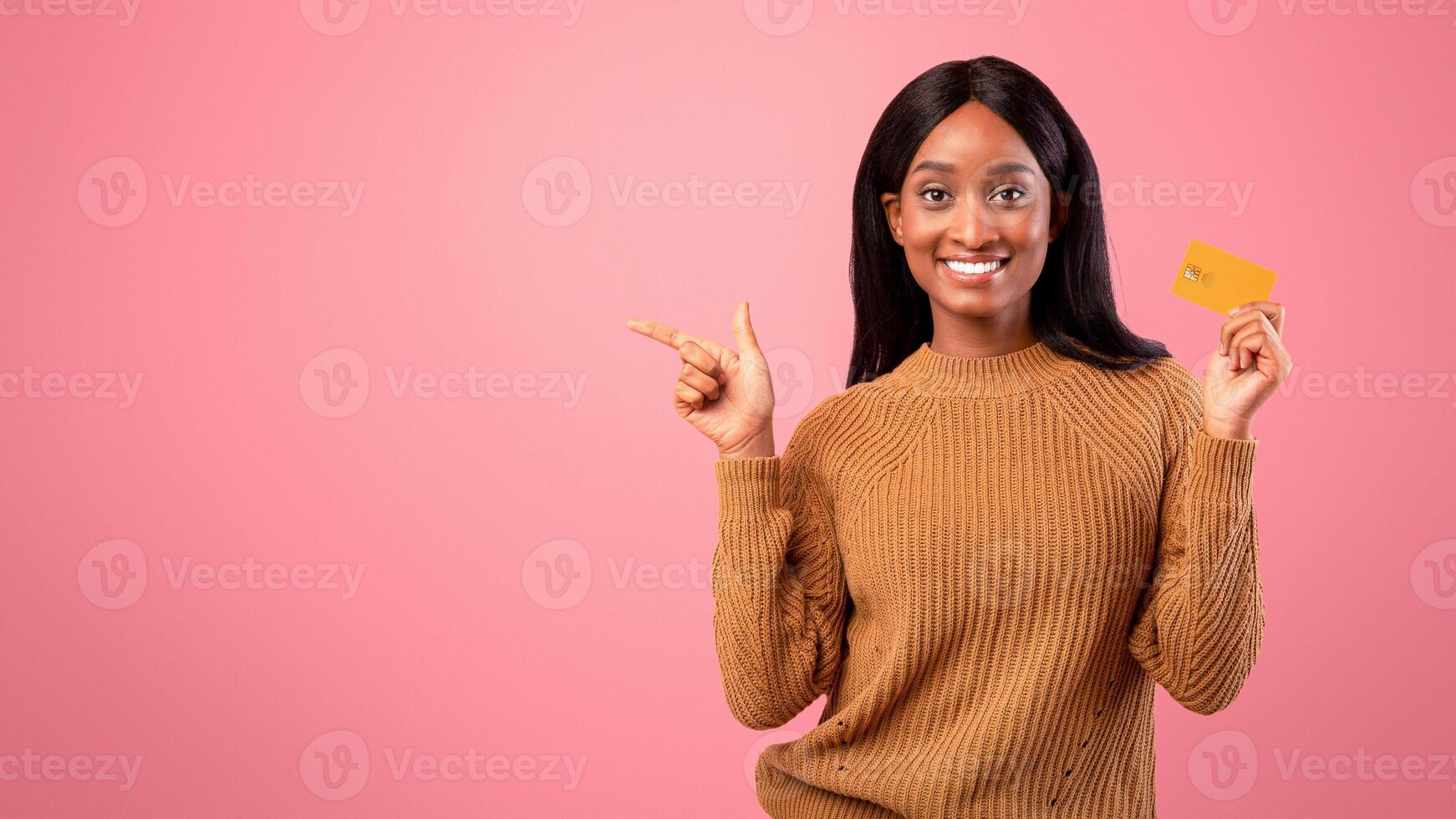 Happy black woman with credit card pointing aside at empty space on pink studio background. Positive female client promoting bank services for contactless payments, panorama photo