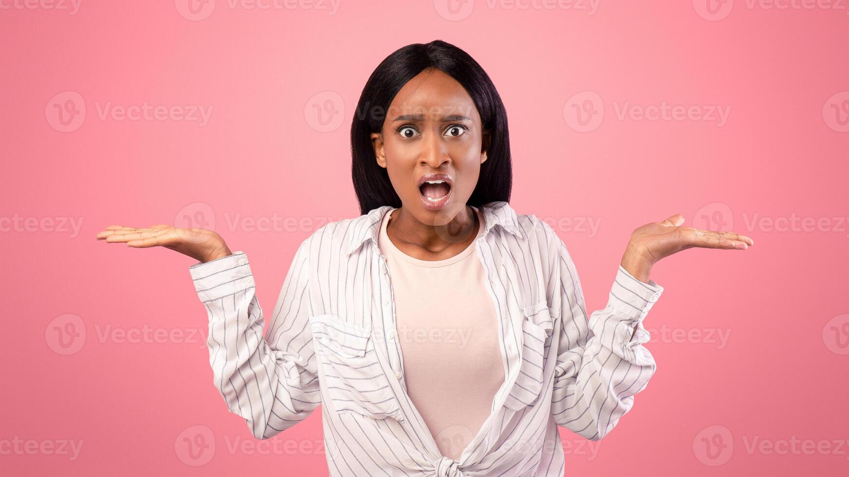 Annoyed African American woman gesturing I DON'T KNOW, shrugging her shoulders on pink studio background. Puzzled black lady cannot understand something, unsure about making choice photo