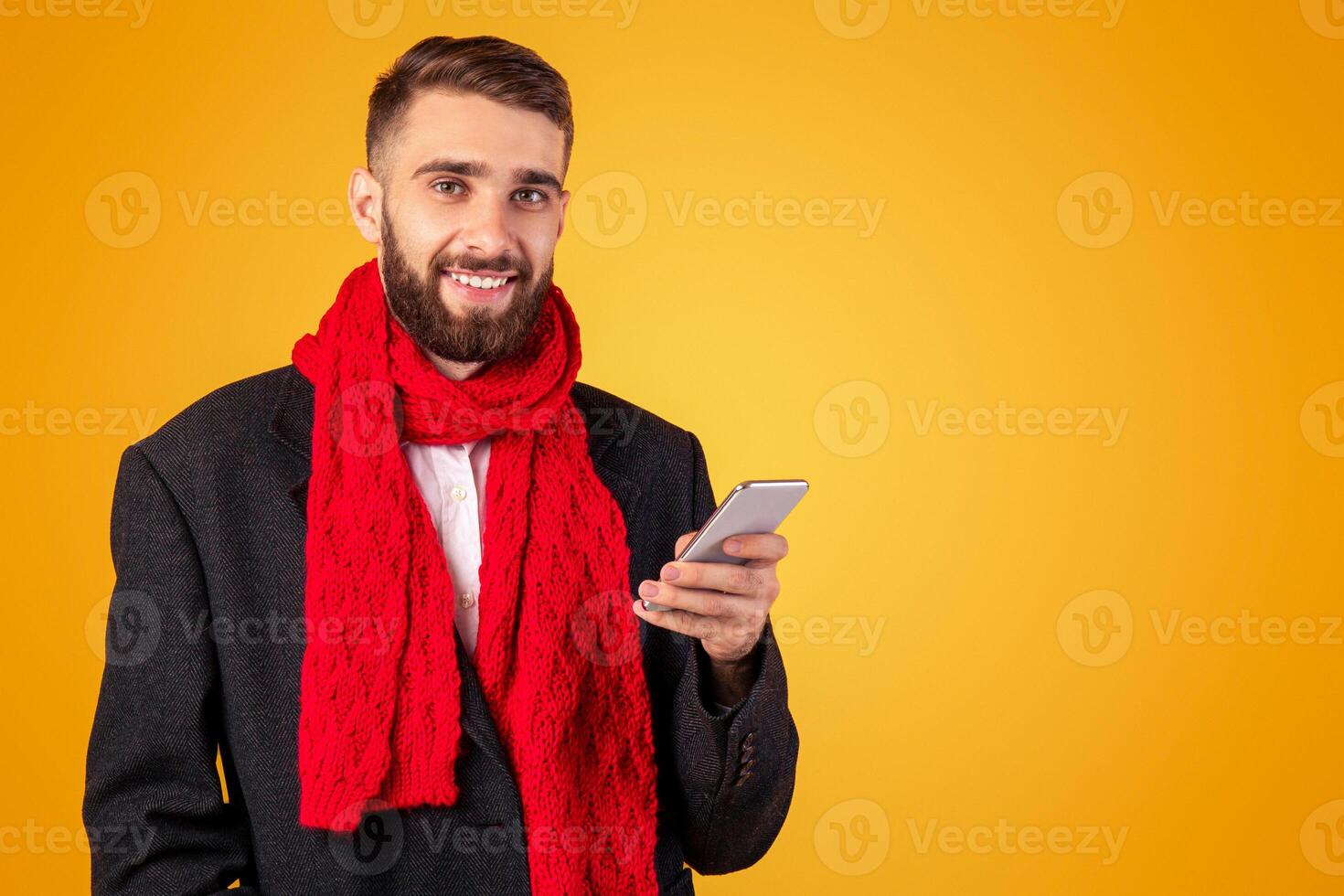Handsome businessman in elegant jacket and red scarf using mobile phone over orange studio background. Positive young guy searching web, watching news or participating in online conference photo