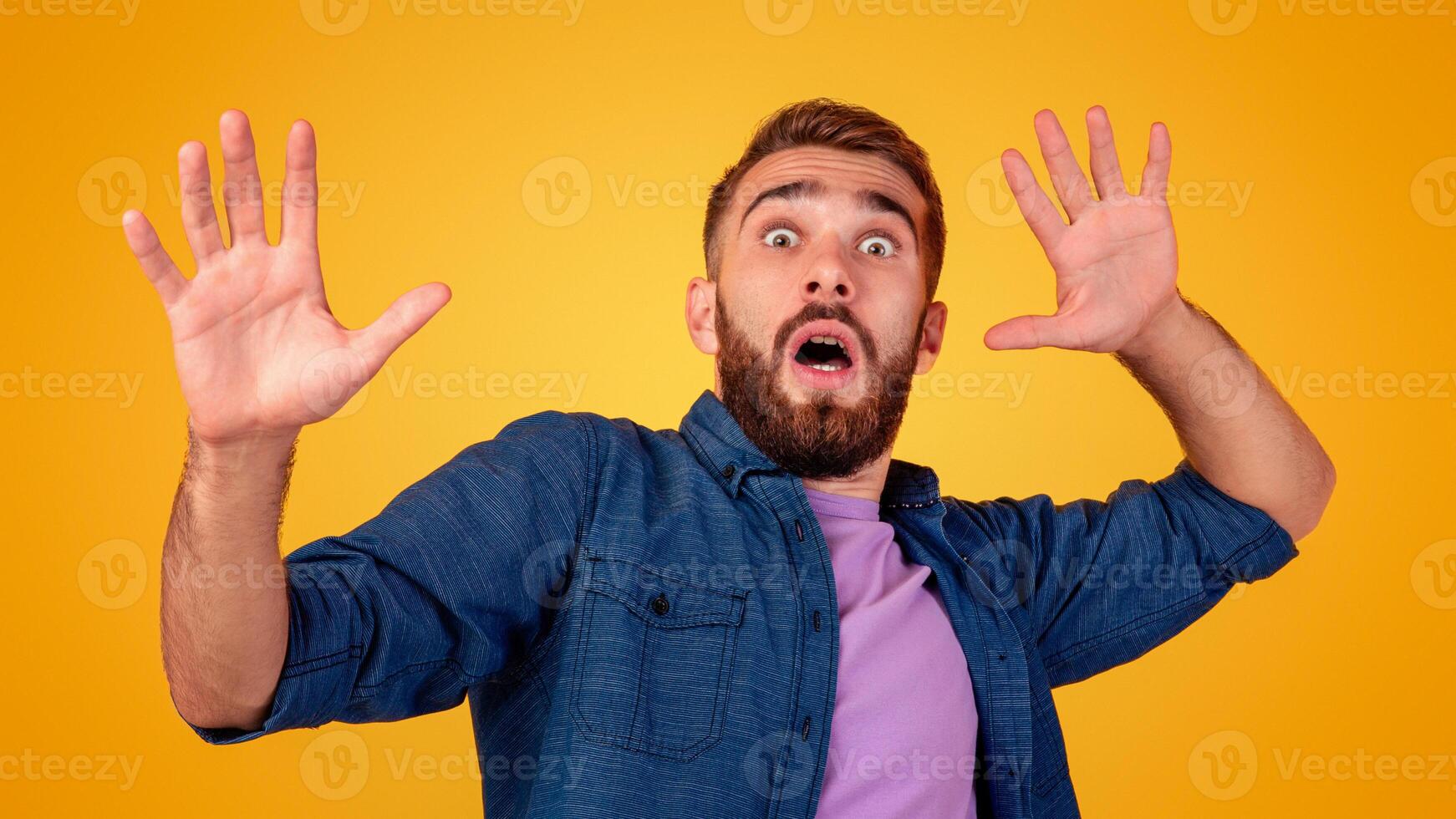 Scared Caucasian man making frightened gesture with his hands, trying to defend himself on orange studio background. Fearful young guy opening his mouth in shock, overwhelmed with emotions photo