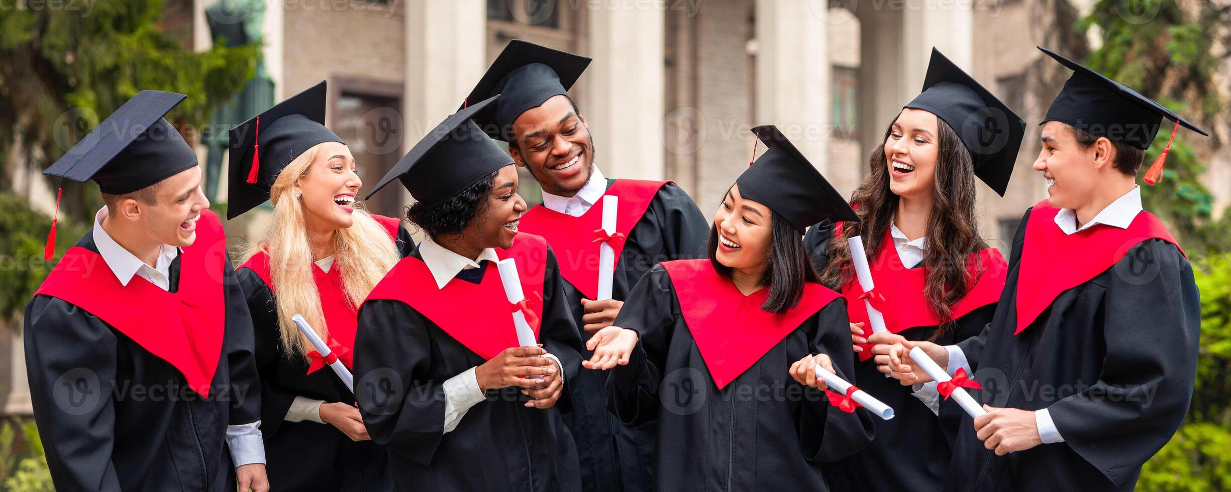 Happy graduates international group of young men and women in graduation costumes holding diplomas, looking at each other and chatting, having ceremony, university campus background photo