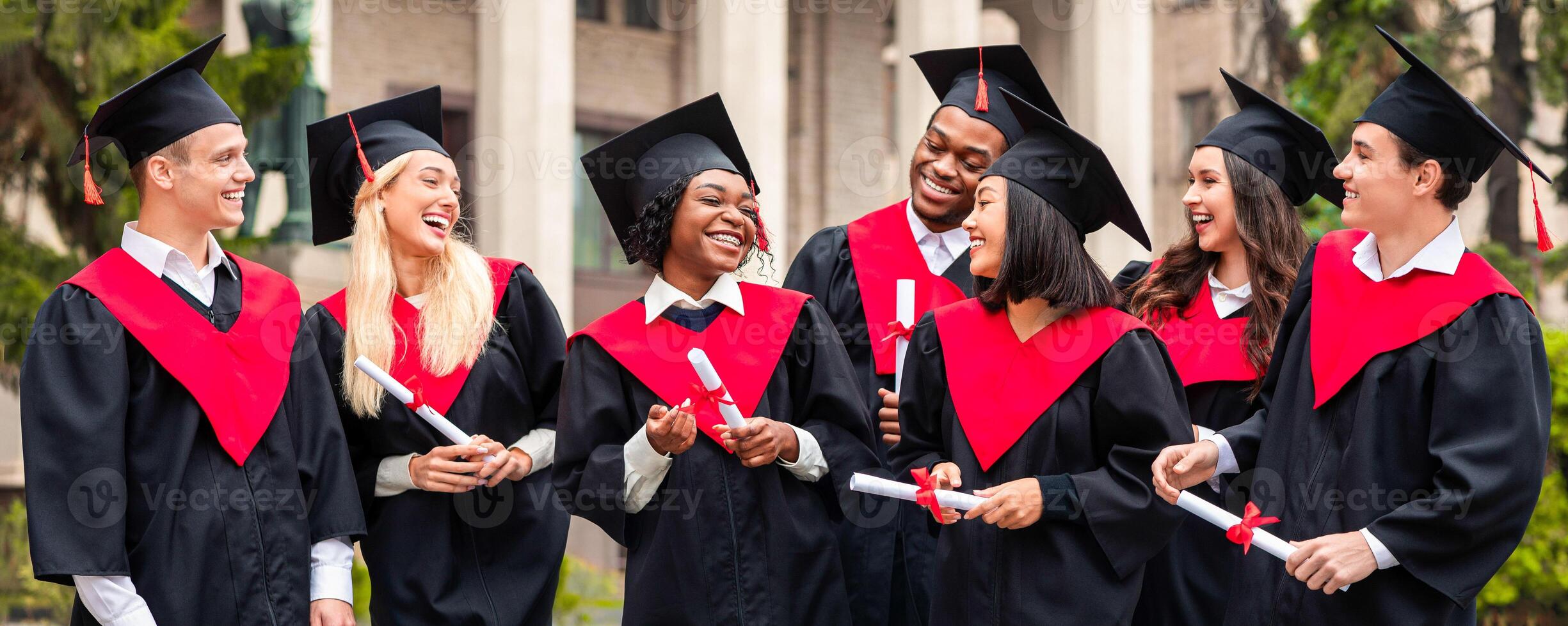 Happy graduates international group of young men and women in graduation costumes holding diplomas, looking at each other and laughing, celebrating success, university campus background photo