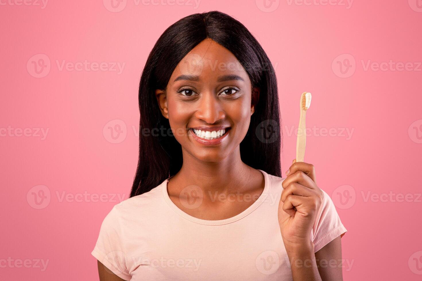 Portrait of beautiful African American woman with radiant smile holding wooden toothbrush with paste on pink studio background. Pretty black lady taking care of her teeth, doing morning routine photo