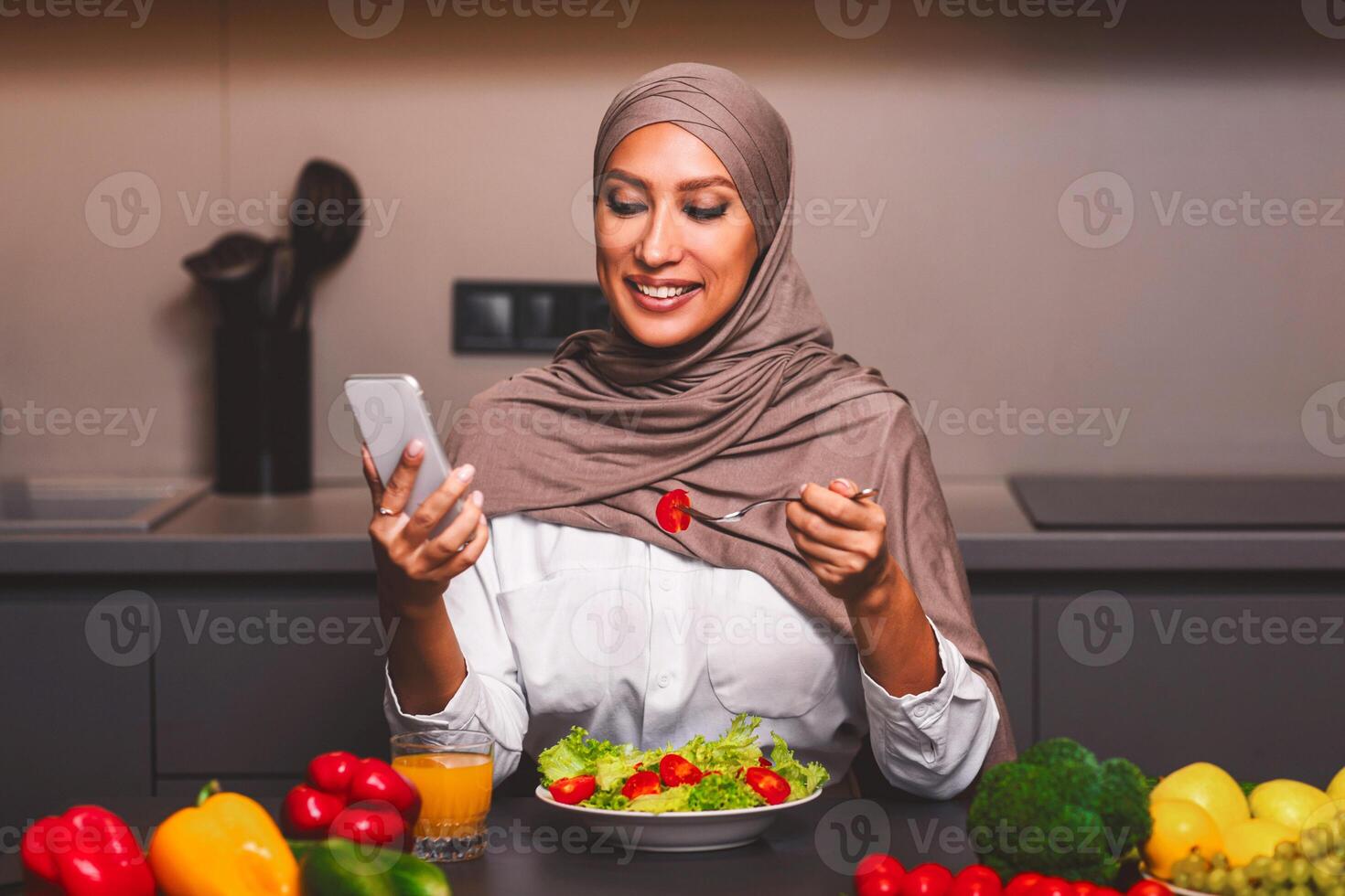 Happy Muslim Woman Using Smartphone Eating Vegetable Salad Having Lunch Sitting In Kitchen At Home. Dieting Mobile Application, Healthy Nutrition And Food, Weight Loss Recipes App For Phone photo