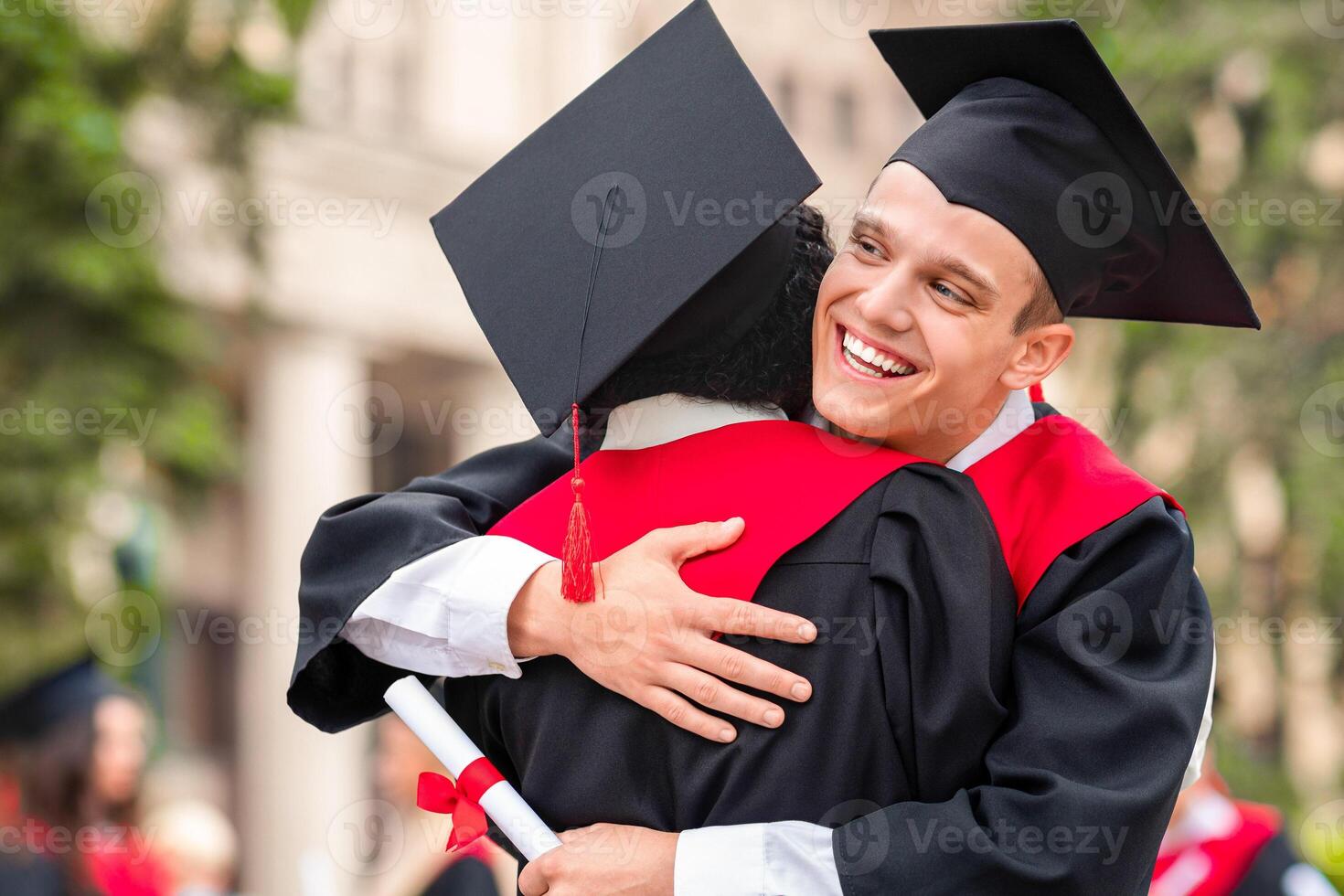 Happy guys international students greeting each other with successful graduation, two young men in graduation robes holding diplomas and hugging, university campus background, closeup photo