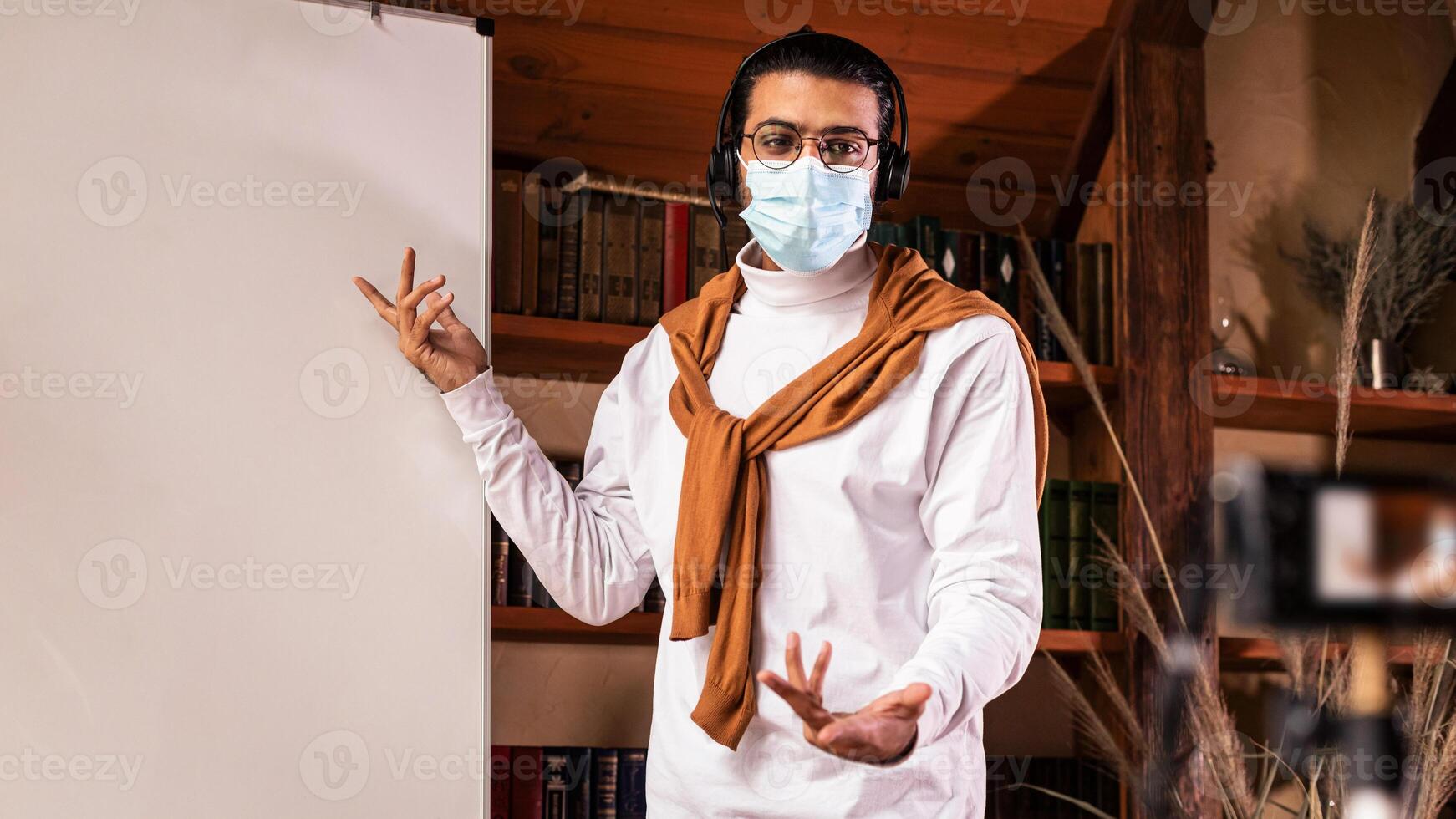 Indian Teacher In Face Mask Making Lecture For Educational Blog Teaching English Language Online, Pointing At Blackboard Explaining Grammar Standing Indoor. Distance Teaching And Education photo