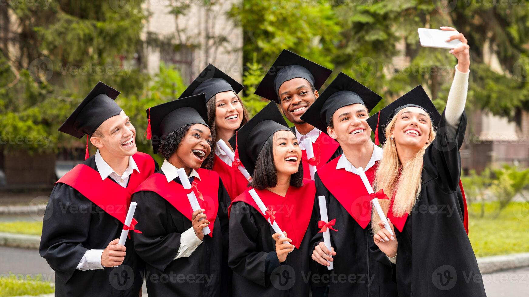 Happy graduates international group of students taking selfie together at university campus, using mobile phone. Cheerful students wearing graduation clothes and holding diplomas photo