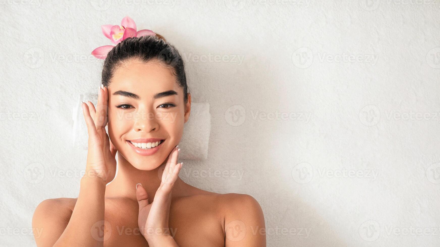 Young Asian Woman Lying On Massage Table, Enjoying Her Soft Smooth Skin After Facial Beauty Treatments In Spa Salon, Having Wellness Day At Cosmetologic Cabinet, Smiling At Camera, Top View photo
