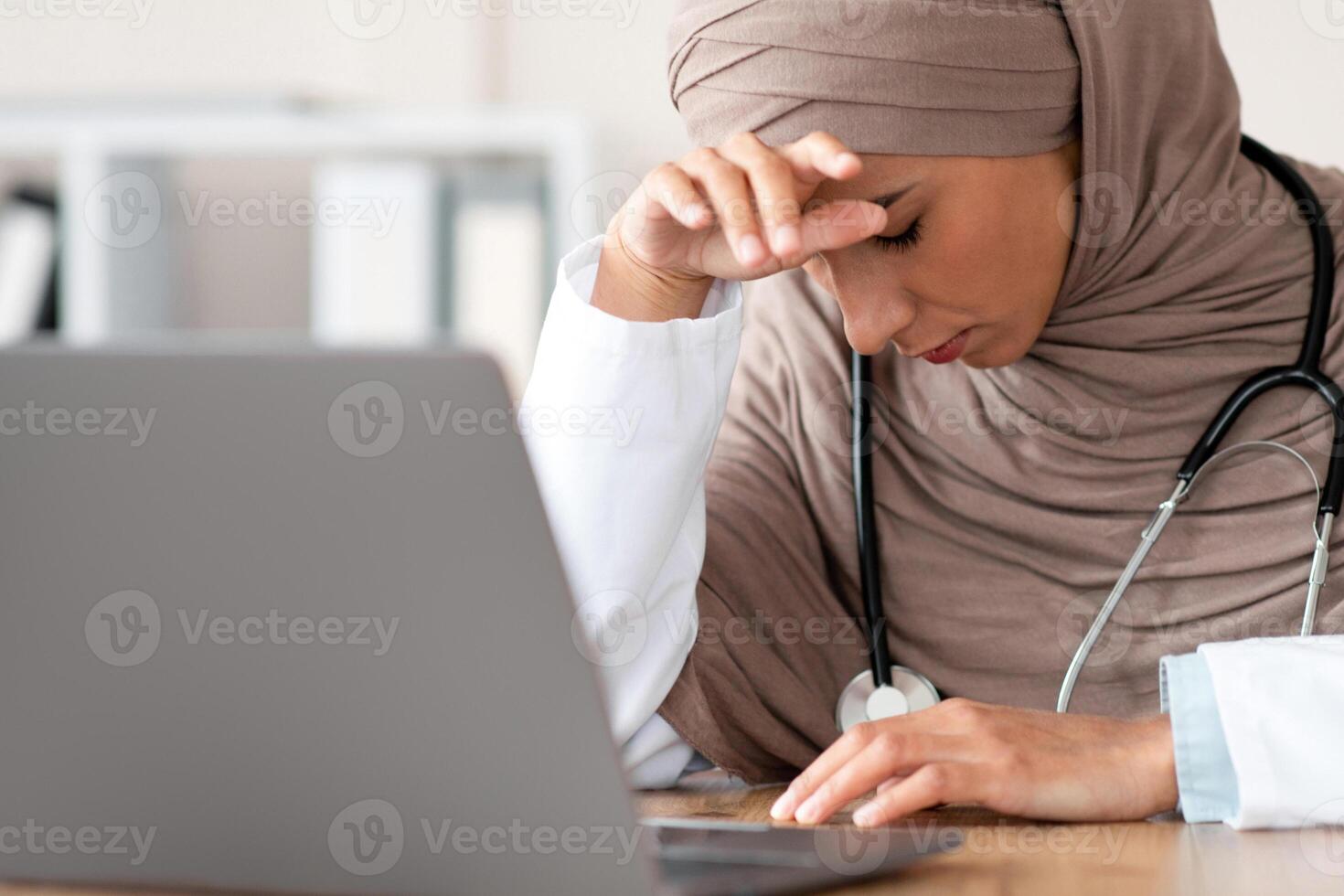 Exhausted muslim woman doctor in hijab working in the clinic, sitting at her workdesk in front of laptop, leaning on her hand, need for break, copy space. Tired lady in hijab physician having headache photo
