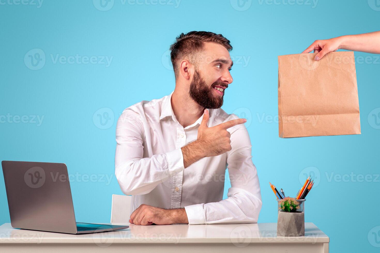 Online food delivery during coronavirus outbreak concept. Happy young guy ordering his takeaway meal to office during lunchtime, sitting at desk with laptop, blue studio background photo