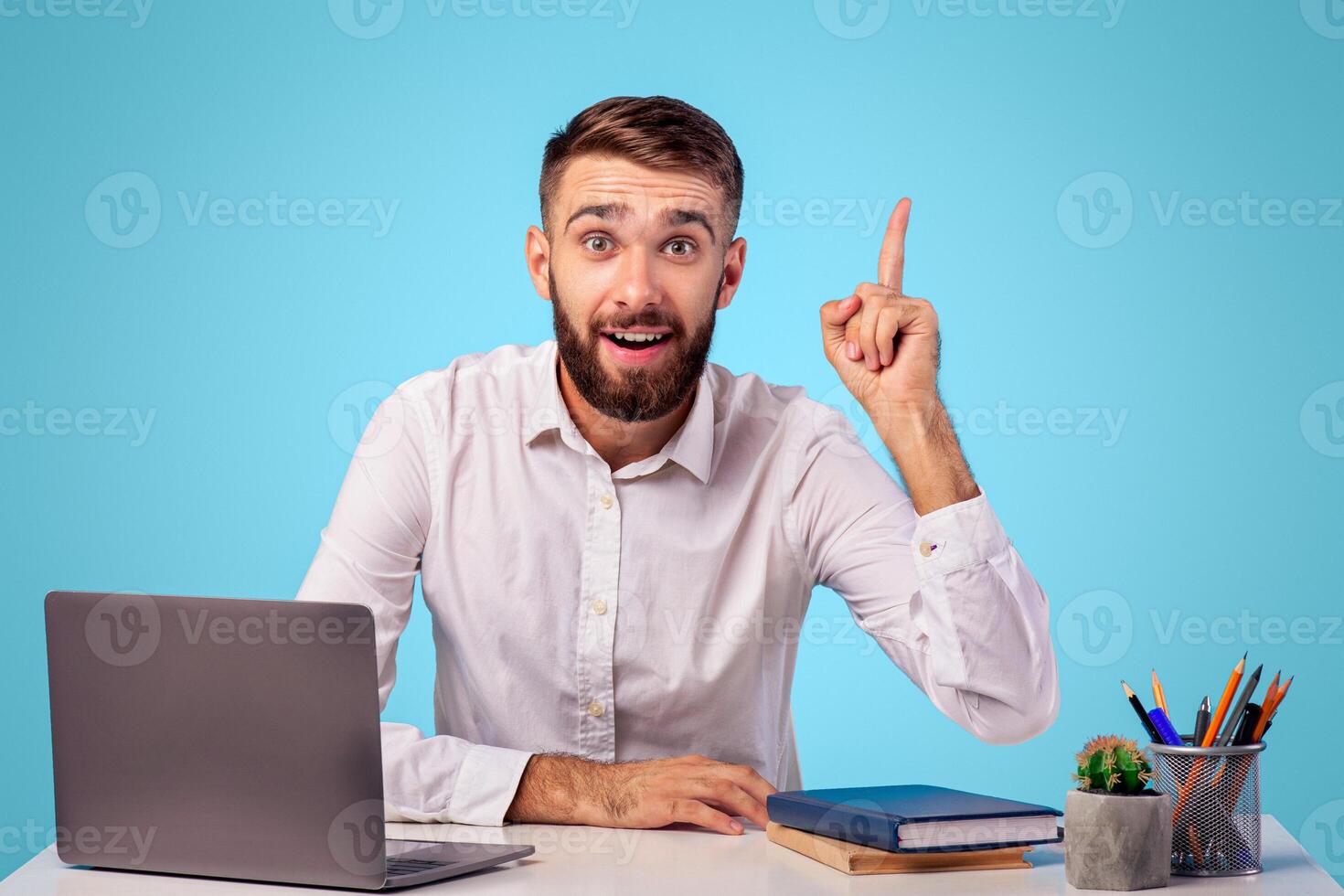 Millennial Caucasian office worker having creative idea, gesturing eureka at workplace on blue studio background. Young man finding solution to problem, sitting at his desk with laptop photo