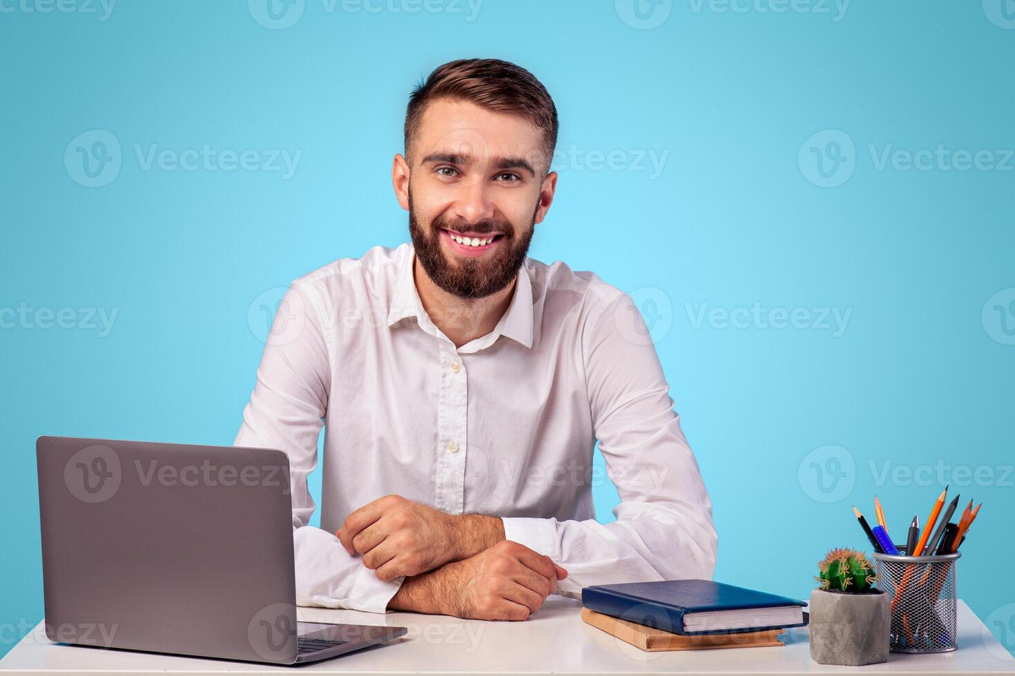 Successful young office worker sitting at desk with laptop computer and smiling on blue studio background. Portrait of happy millennial businessman in formal wear at workplace photo