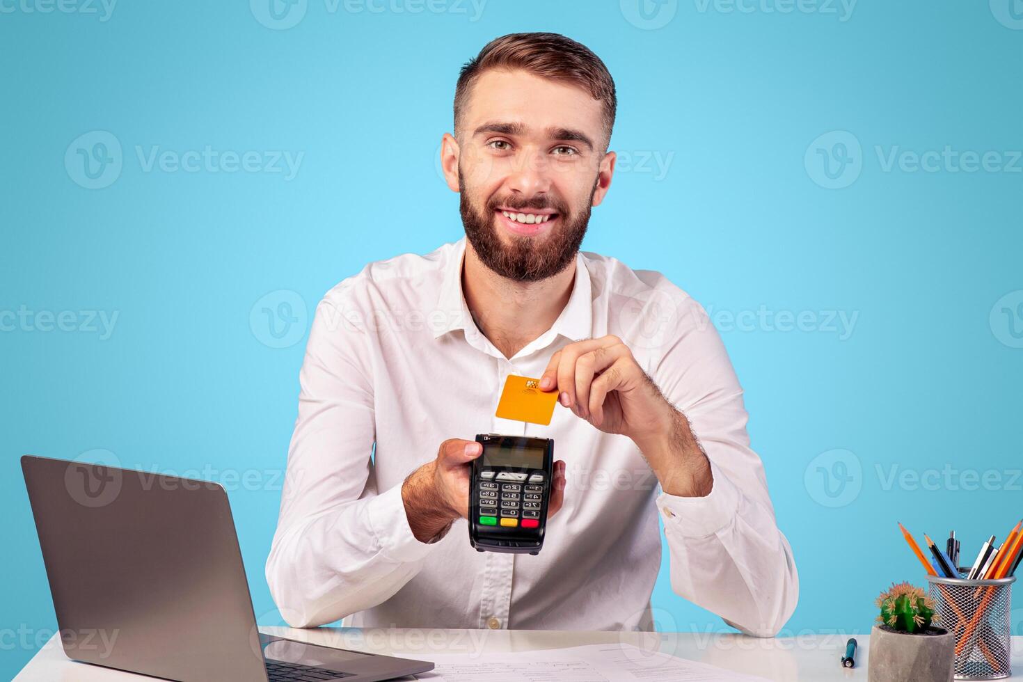 Happy young man sitting at desk in office, holding money terminal and credit card, blue background. Millennial company employee paying contactless, using electronic currency for online shopping photo