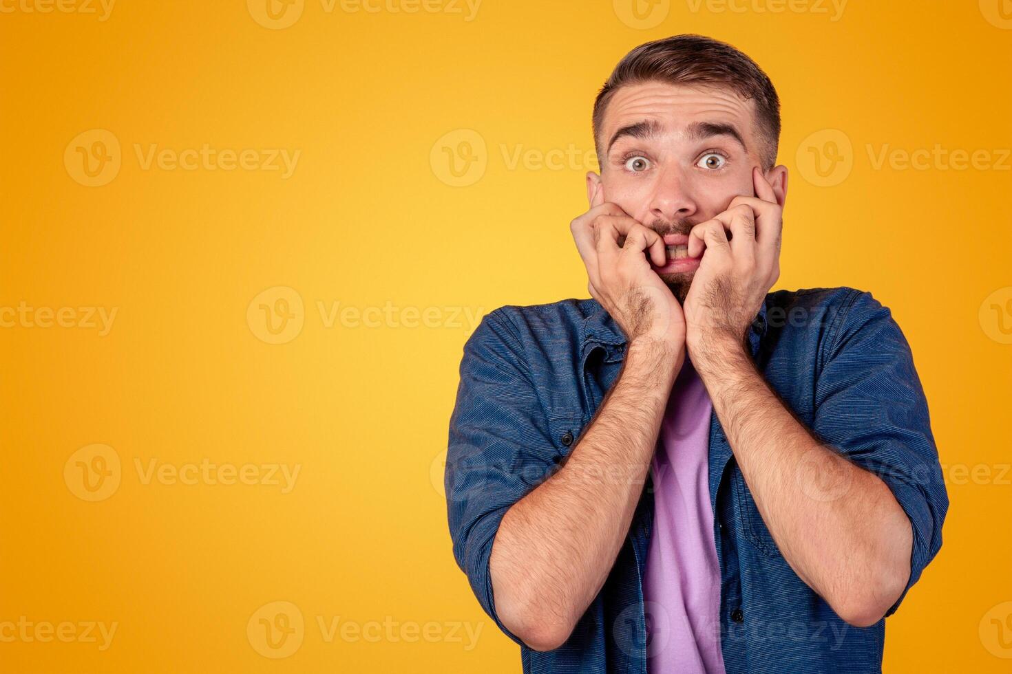 Scared Caucasian guy biting his nails in terror over grey studio background. Young man feeling terrified, suffering from fear or shock, having panic attack. Expressing negative emotions concept photo
