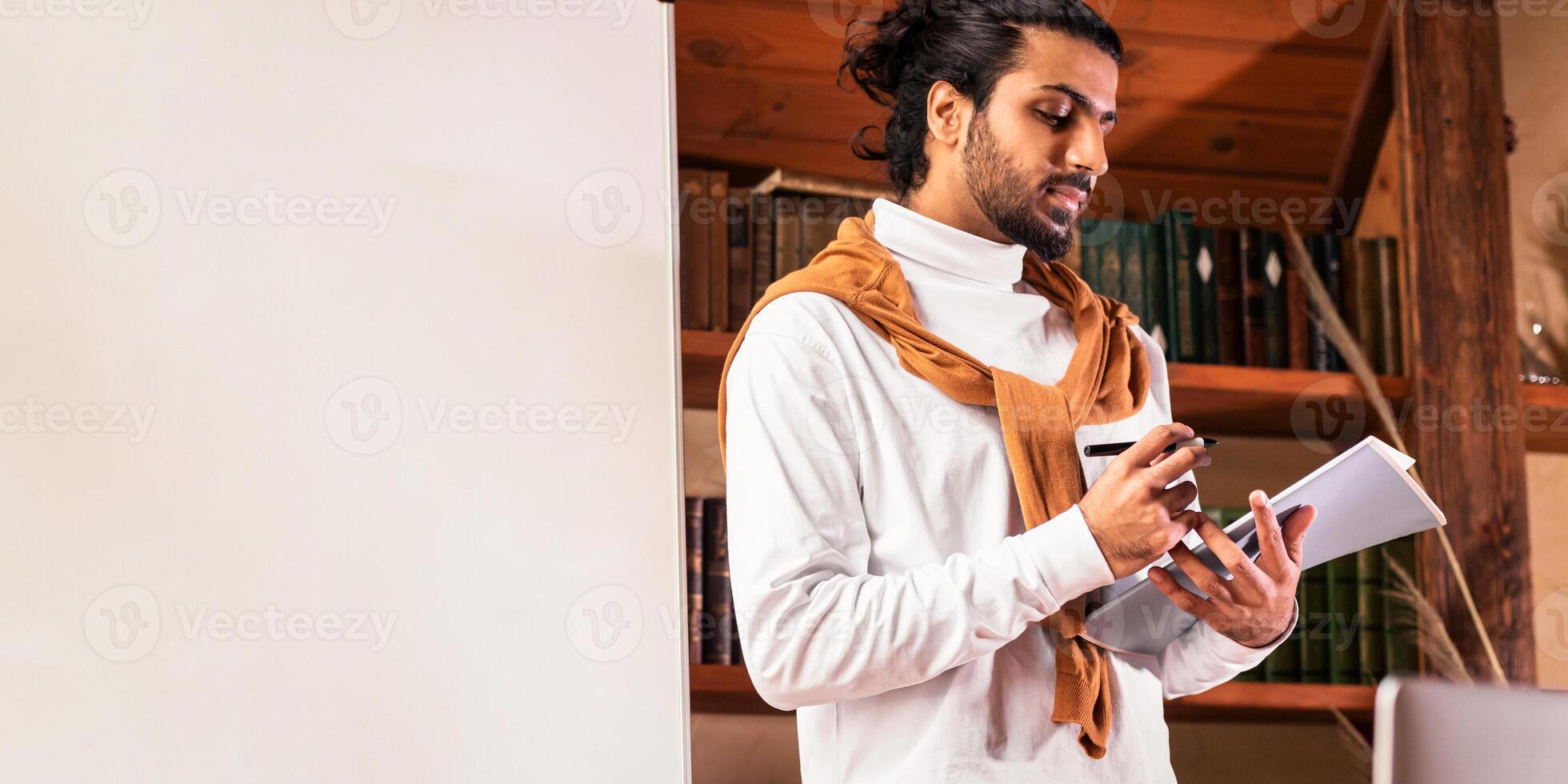 E-Teaching. Indian Teacher Man Standing Near Laptop And Blackboard Having Distant Online Class Indoors. Modern Remote Teaching, Internet Education. Selective Focus photo