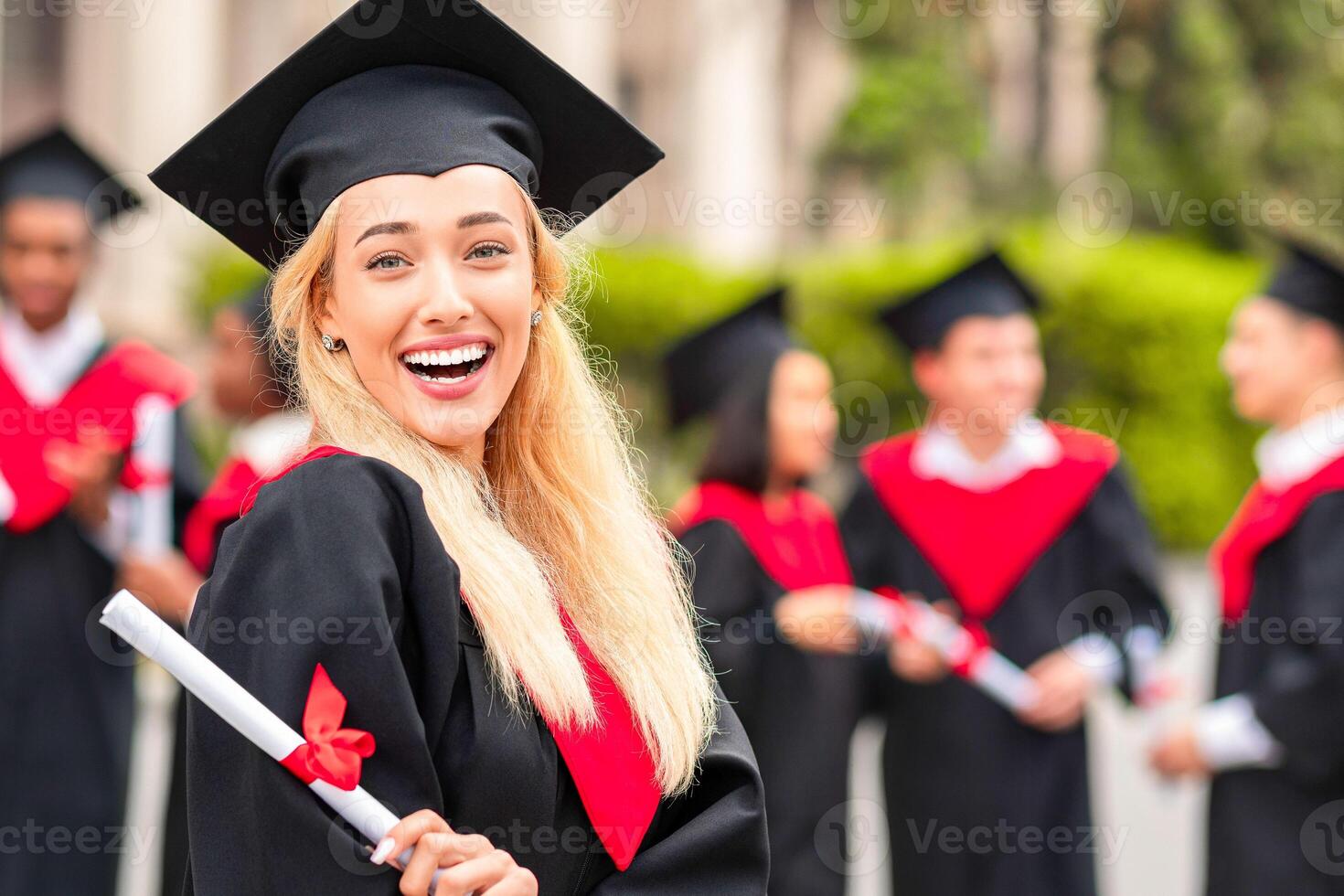 Happy young woman student cheerfully posing while graduation party, wearing graduation robe and hat, holding diploma and smiling at camera, international group of students on background photo