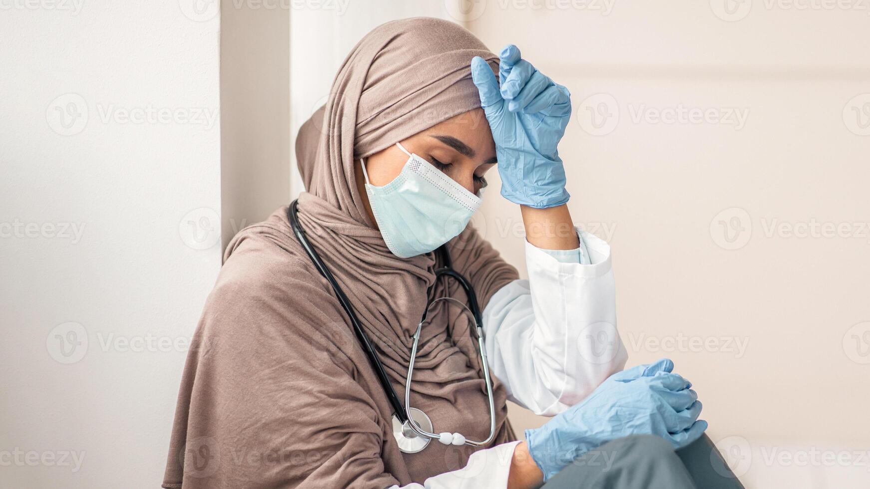 Upset woman in hijab doctor having difficult day at clinic, wearing protective gloves and face mask, sitting next to wall in her office and resting, empty space. Sad female doctor feeling exhausted photo