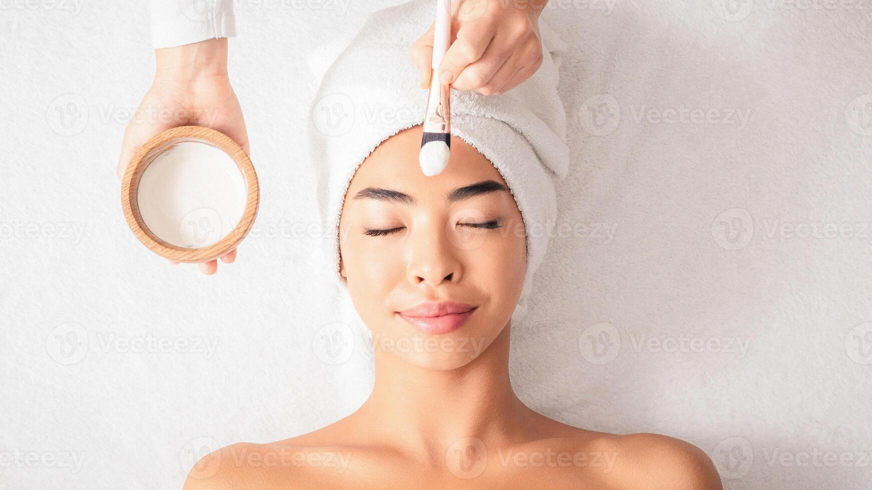 Beauty Treatments. Cosmetologist applying clay mask on asian woman face at spa salon, relaxed korean lady with towel on head enjoying wellness day at beauty center, top view with free space photo
