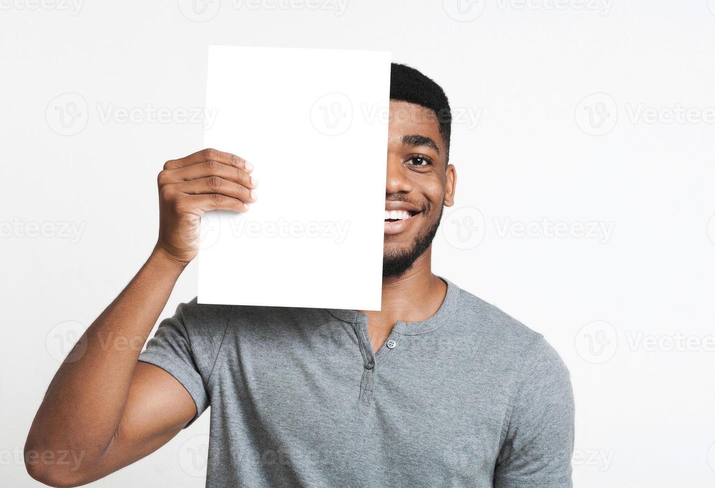 Hi friend. Smiling african-american man covering half of his face with clean sheet of paper, copy space photo