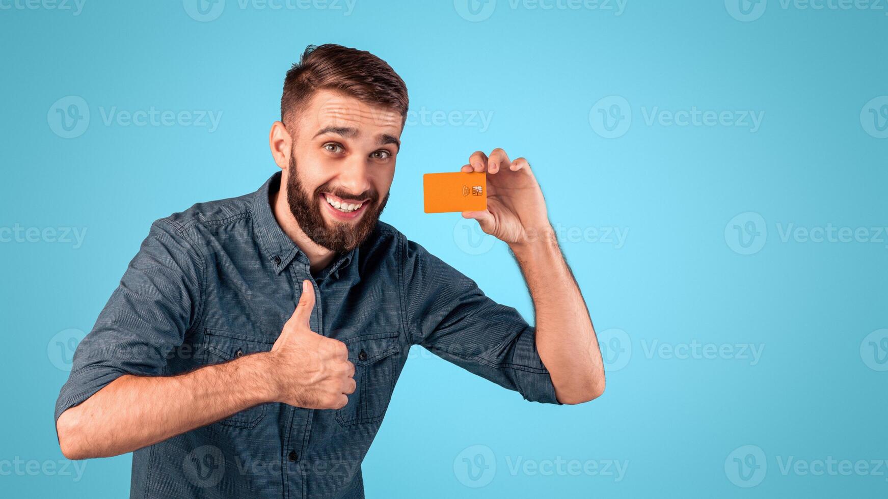 Cheerful young guy with credit card showing thumb up gesture on blue studio background. Satisfied bank client recommending financial services, promoting contactless payment photo