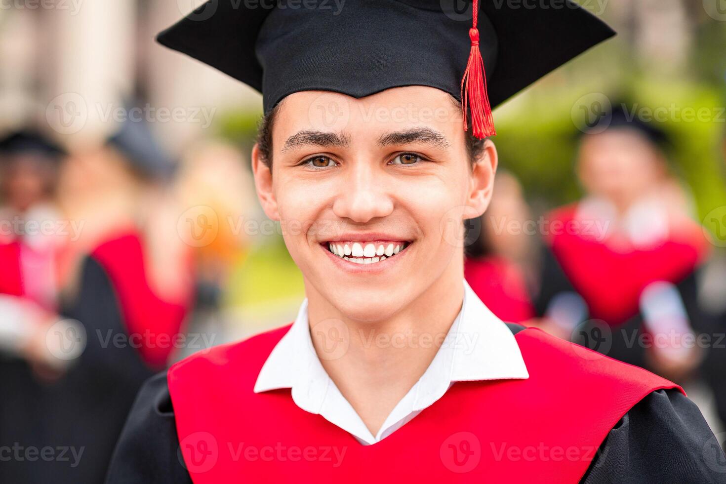Closeup portrait of happy guy student wearing graduating costume. Positive smiling male student celebrating graduation with his friends at university campus, blurred background photo