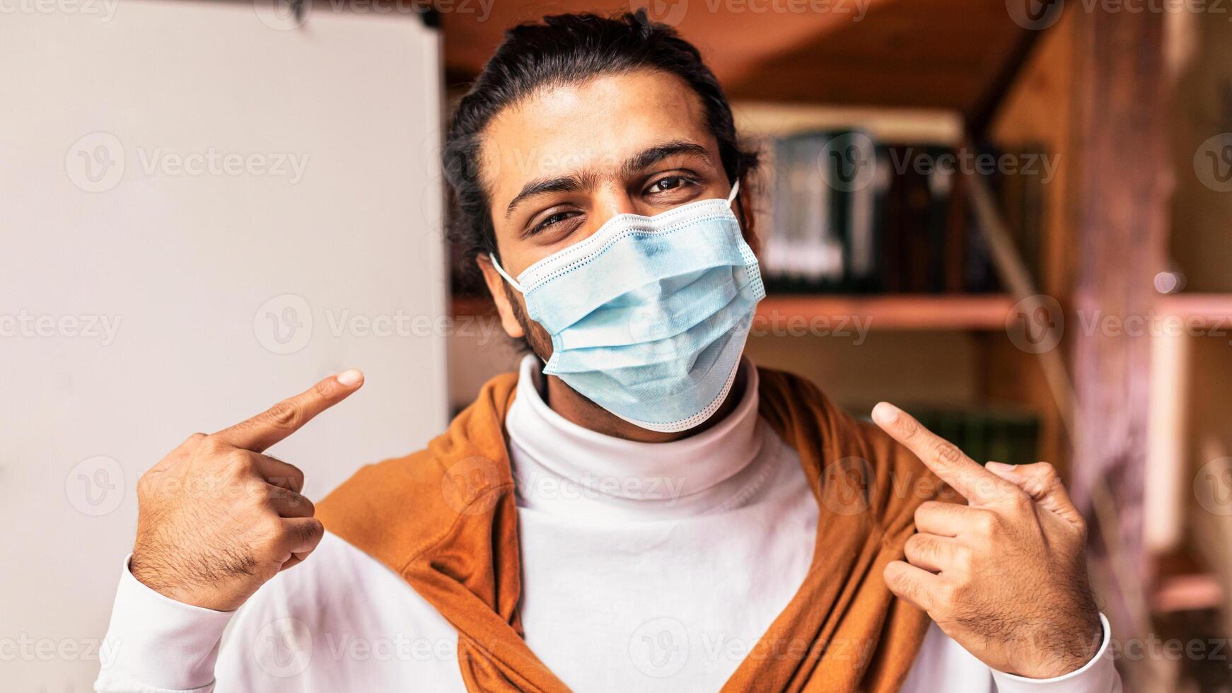 Indian Teacher Pointing At Protective Mask On Face Posing Looking At Camera Near Blackboard Indoor, Announcing Online Distance Learning Quarantine At School And University Due To Coronavirus Outbreak photo