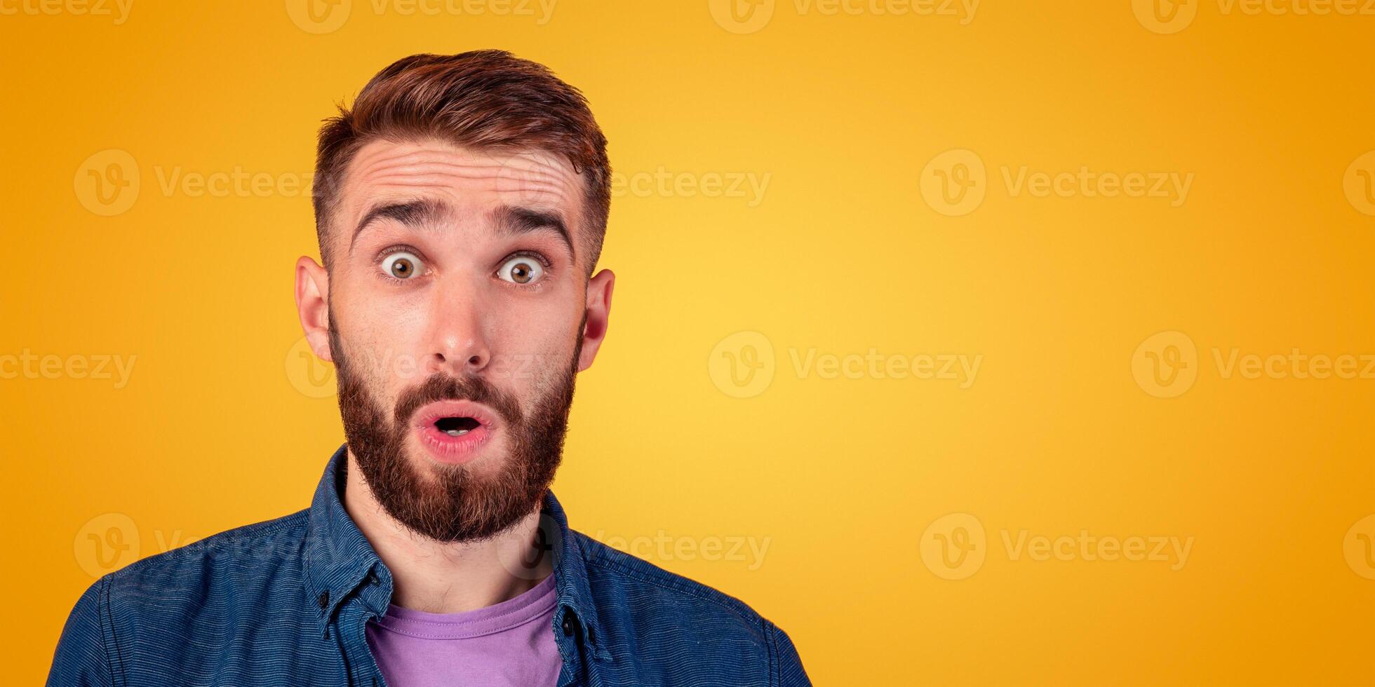 Unbelievable news. Portrait of bearded young guy opening his mouth and eyes in shock, looking at camera on orange studio background. Thunderstruck millennial man feeling astonished photo